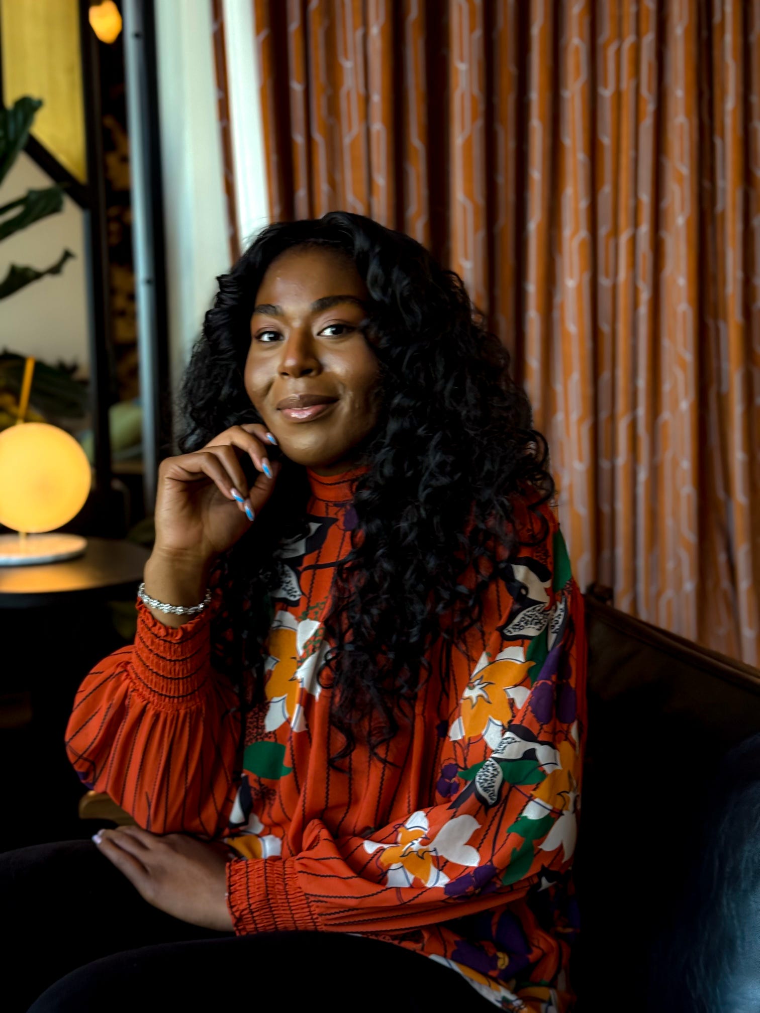 Woman with long curly hair smiling, sitting indoors with curtains and warm lighting behind her.
