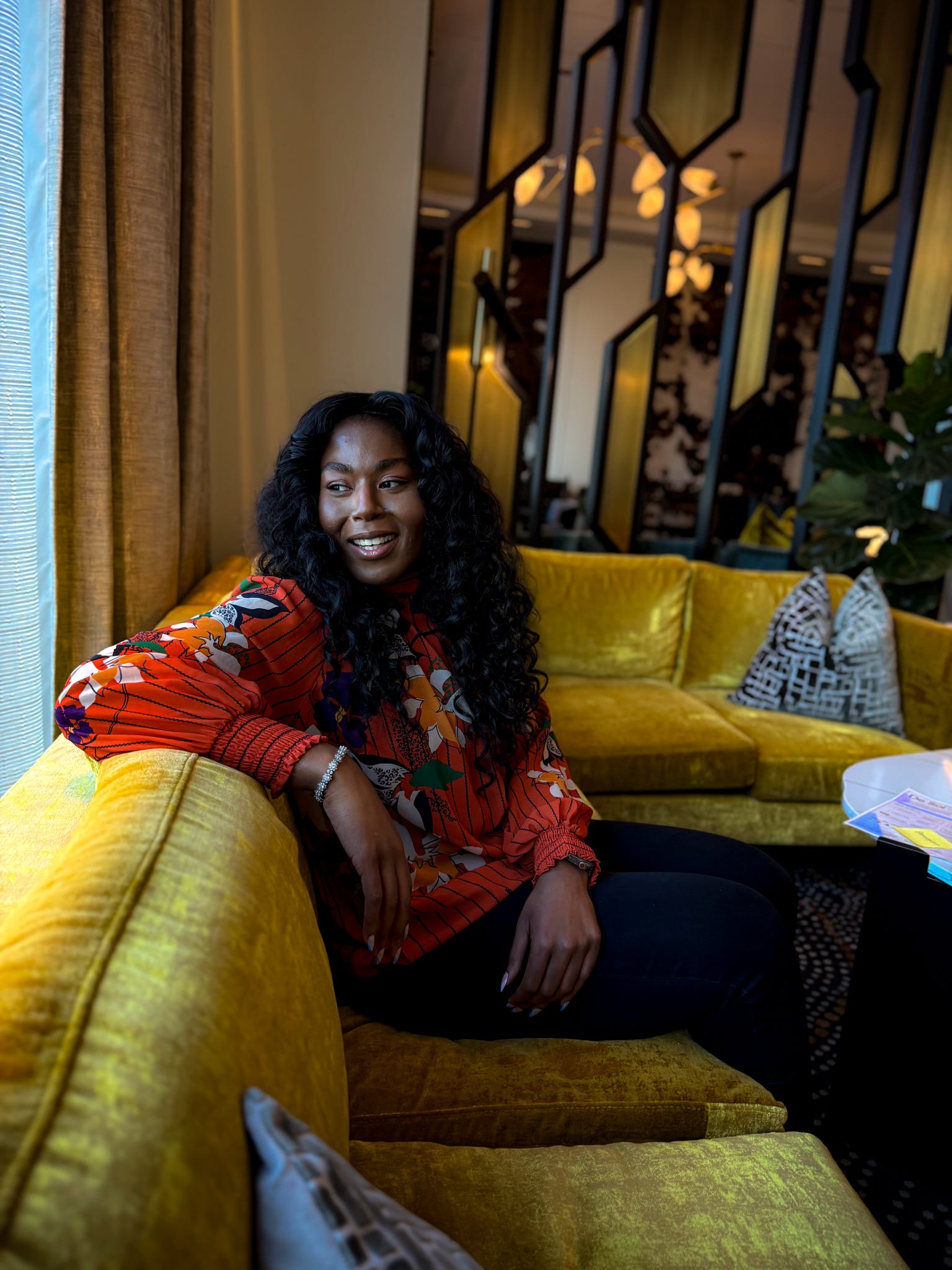 Woman with curly hair smiling, sitting on a yellow sofa in a warmly lit room with decorative elements.