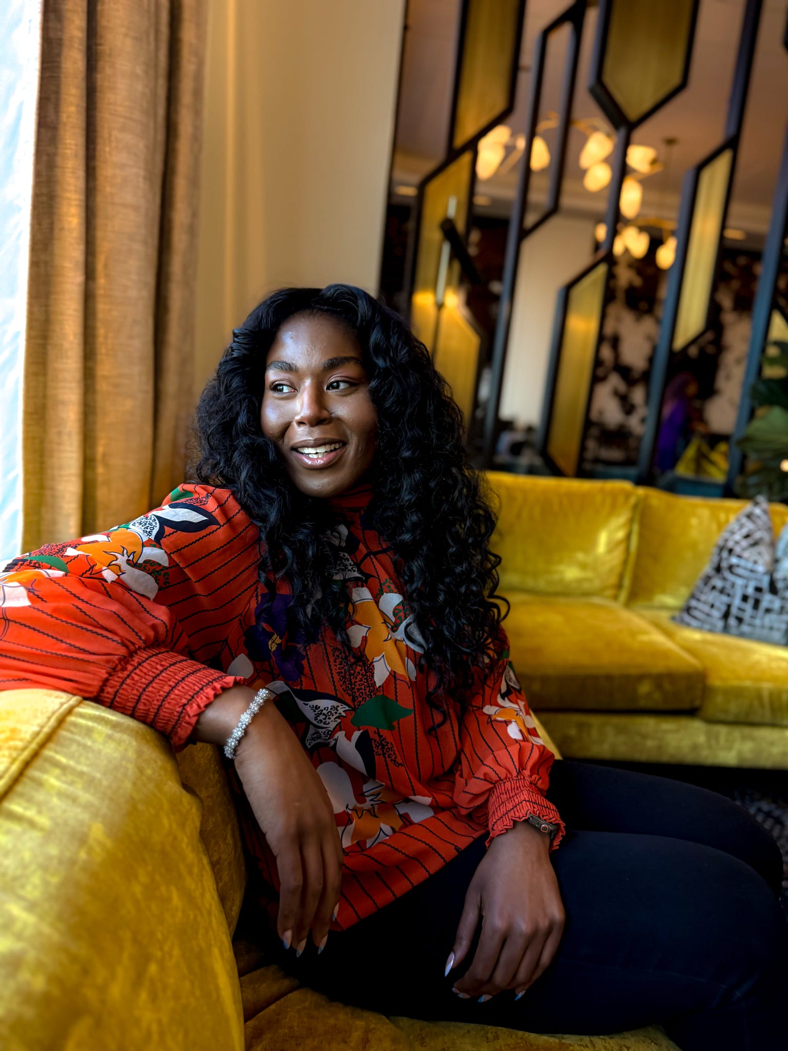Woman with long curly hair sitting on a yellow sofa in a stylish room, smiling, with decorative mirrors and warm lighting.