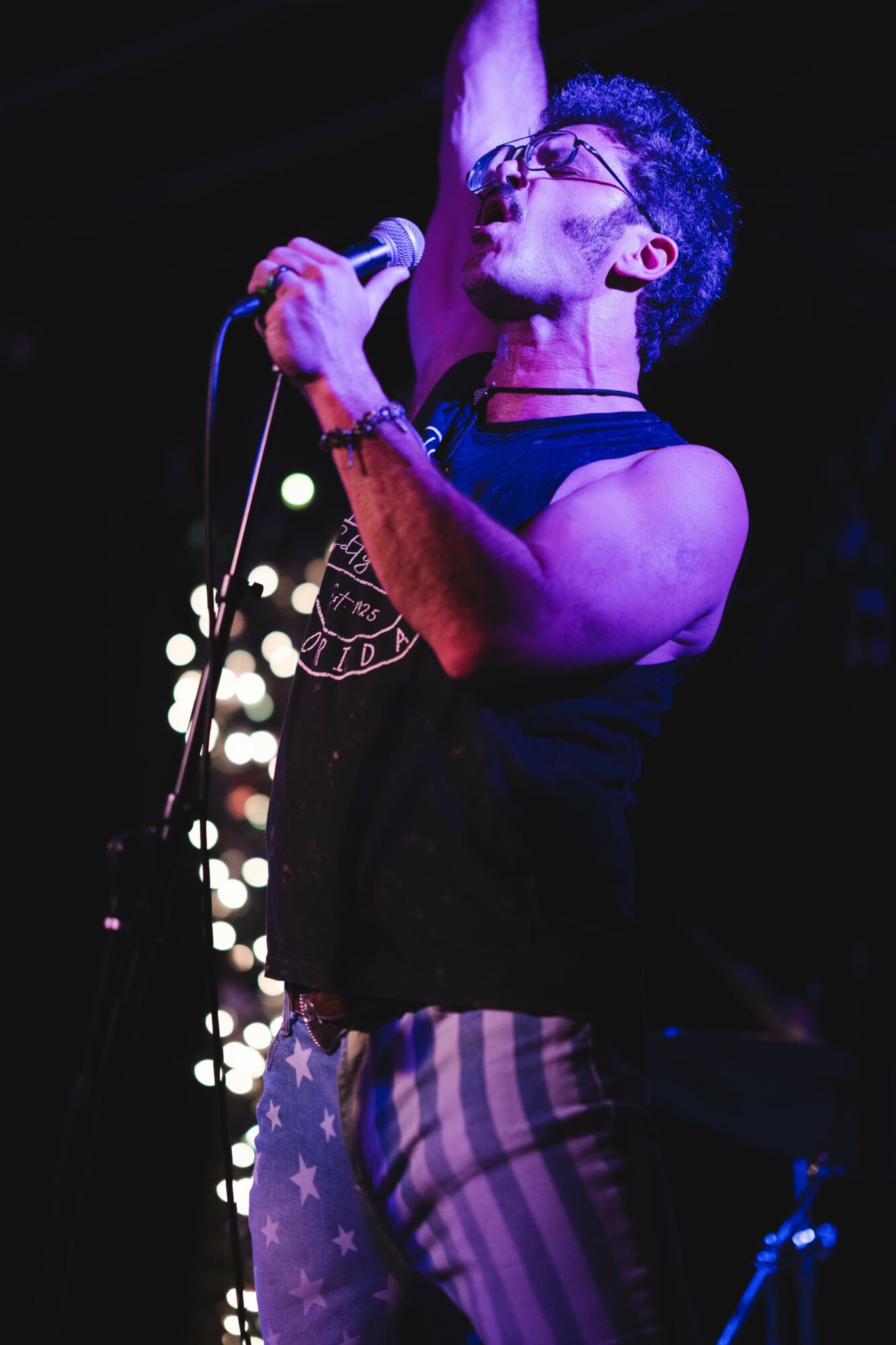 Person singing into a microphone on stage, wearing glasses and striped pants, with stage lights in background.