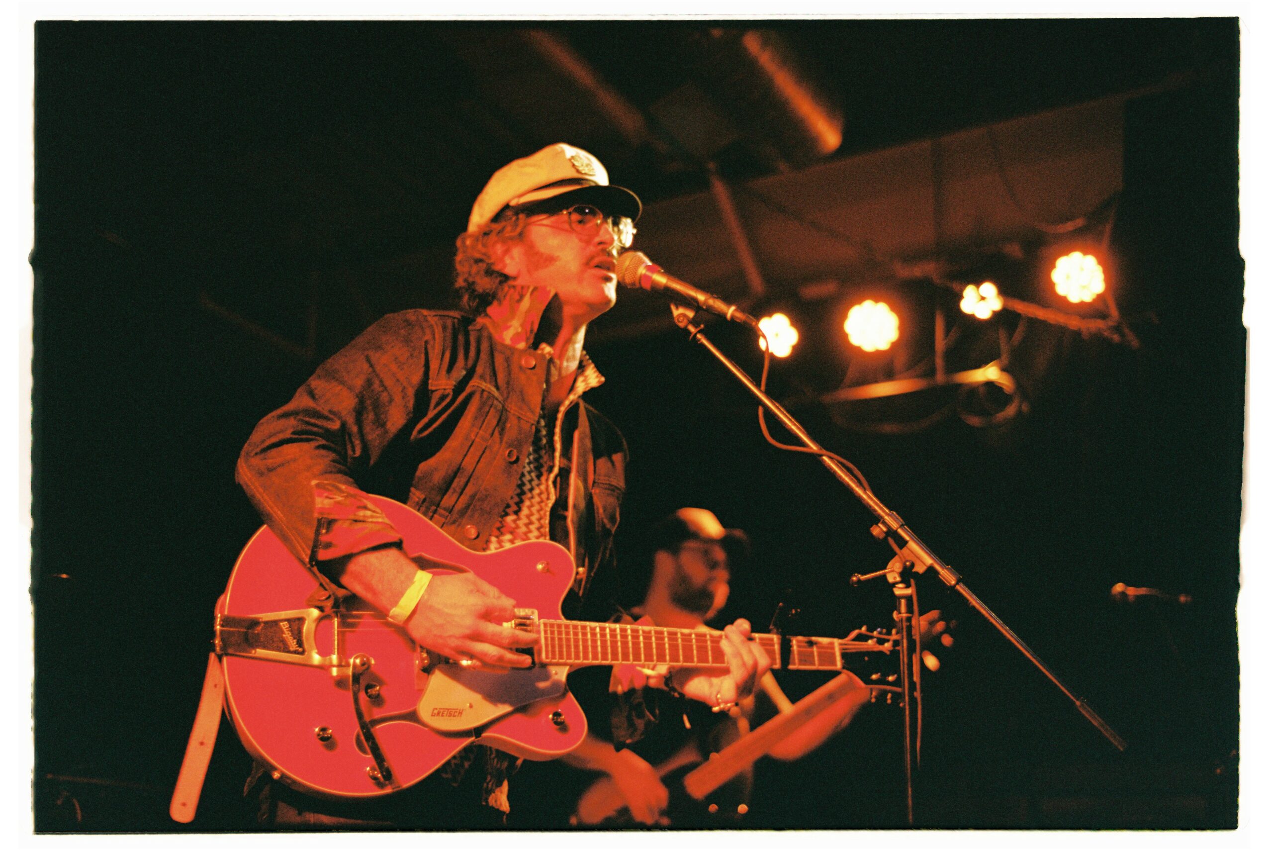 Musician with curly hair, wearing a cap, playing an electric guitar on stage with bright lights behind.