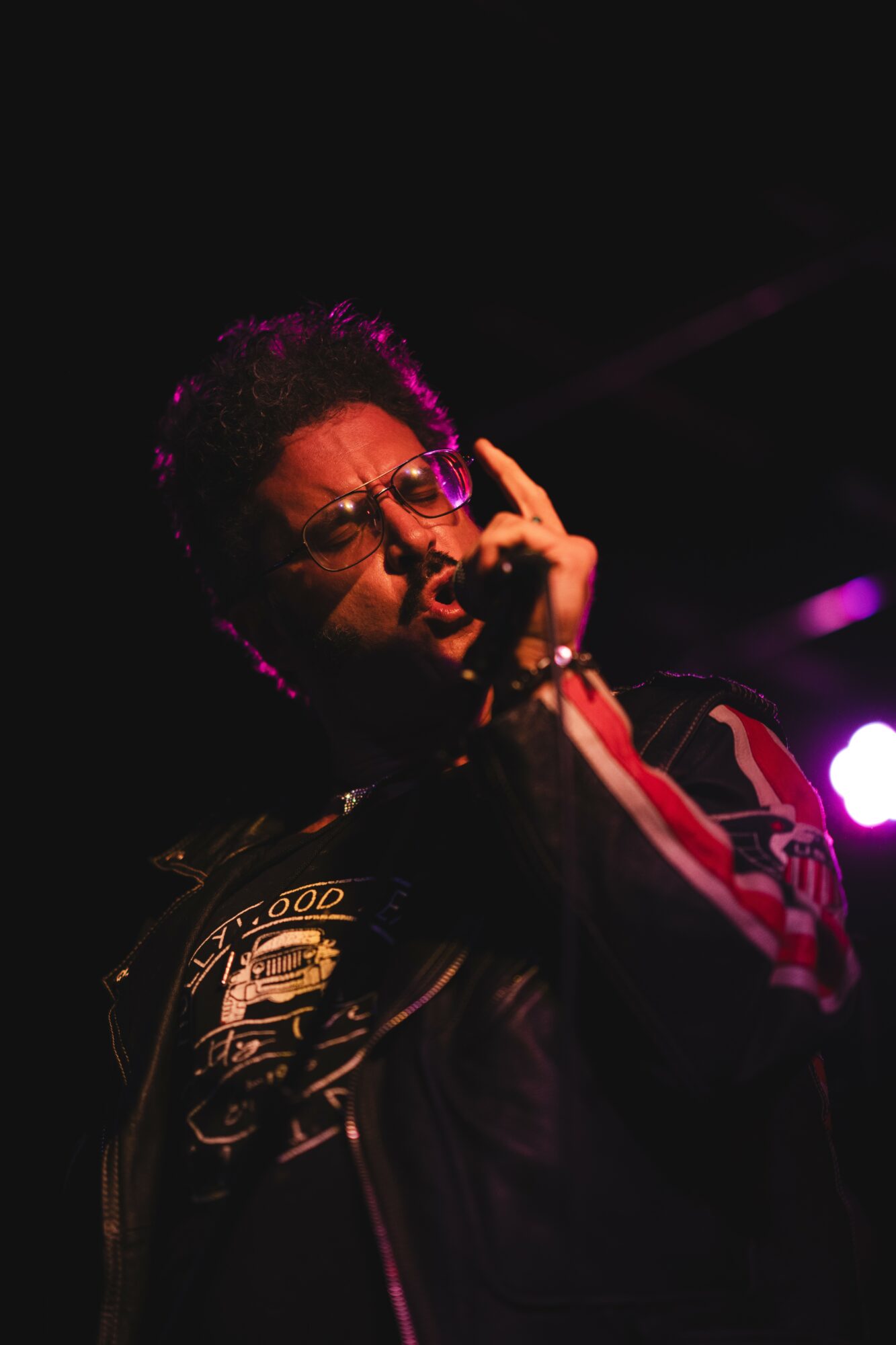 Person with curly hair and glasses making a gesture with their hand, wearing a dark jacket with patches, in a dark setting.