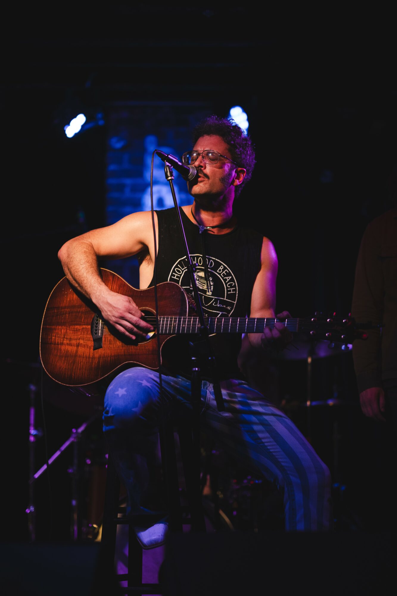 Person playing acoustic guitar and singing into microphone on stage with blue lighting, wearing glasses and sleeveless shirt.