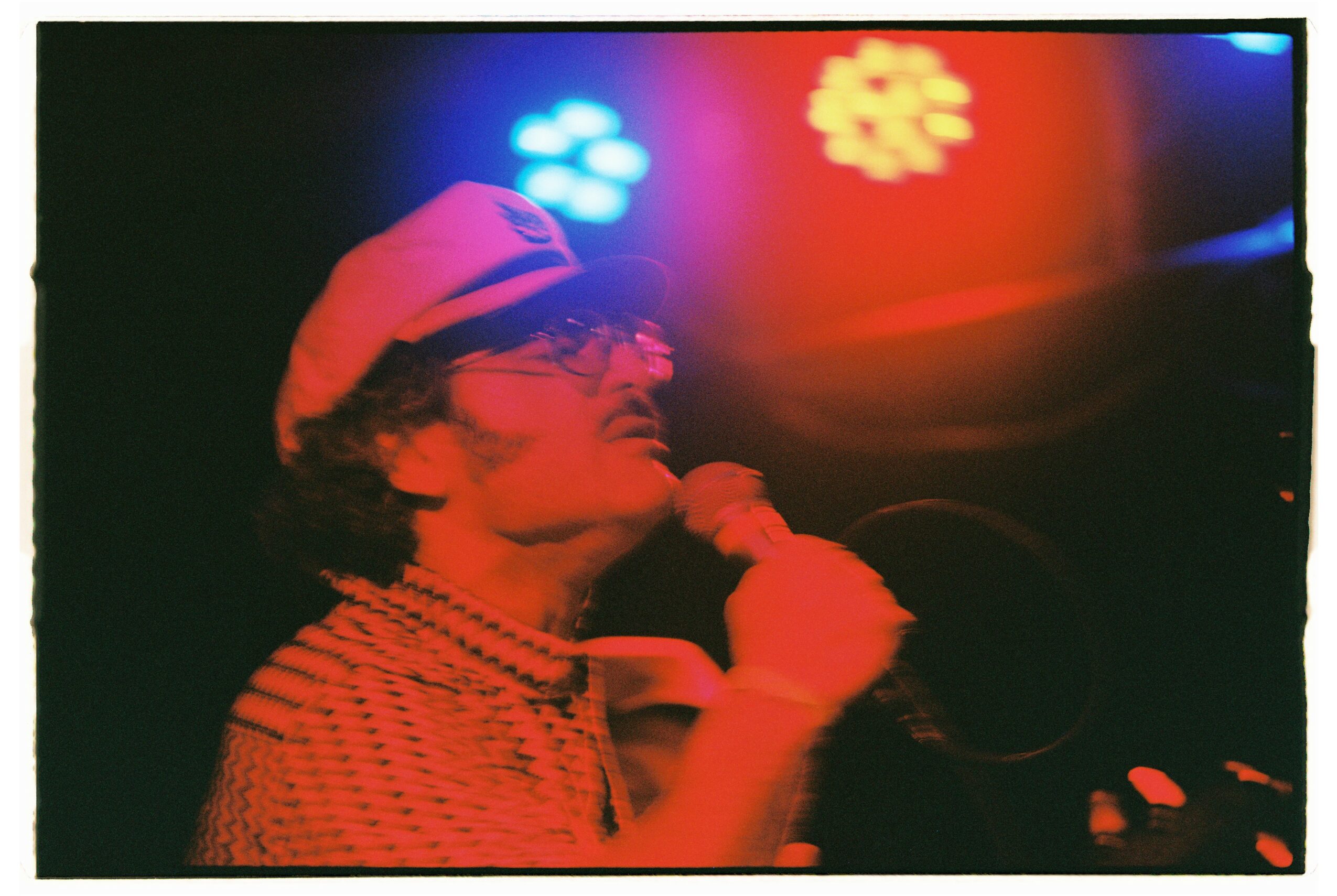 Person wearing a hat and glasses, holding a microphone, with colorful lights in the background.