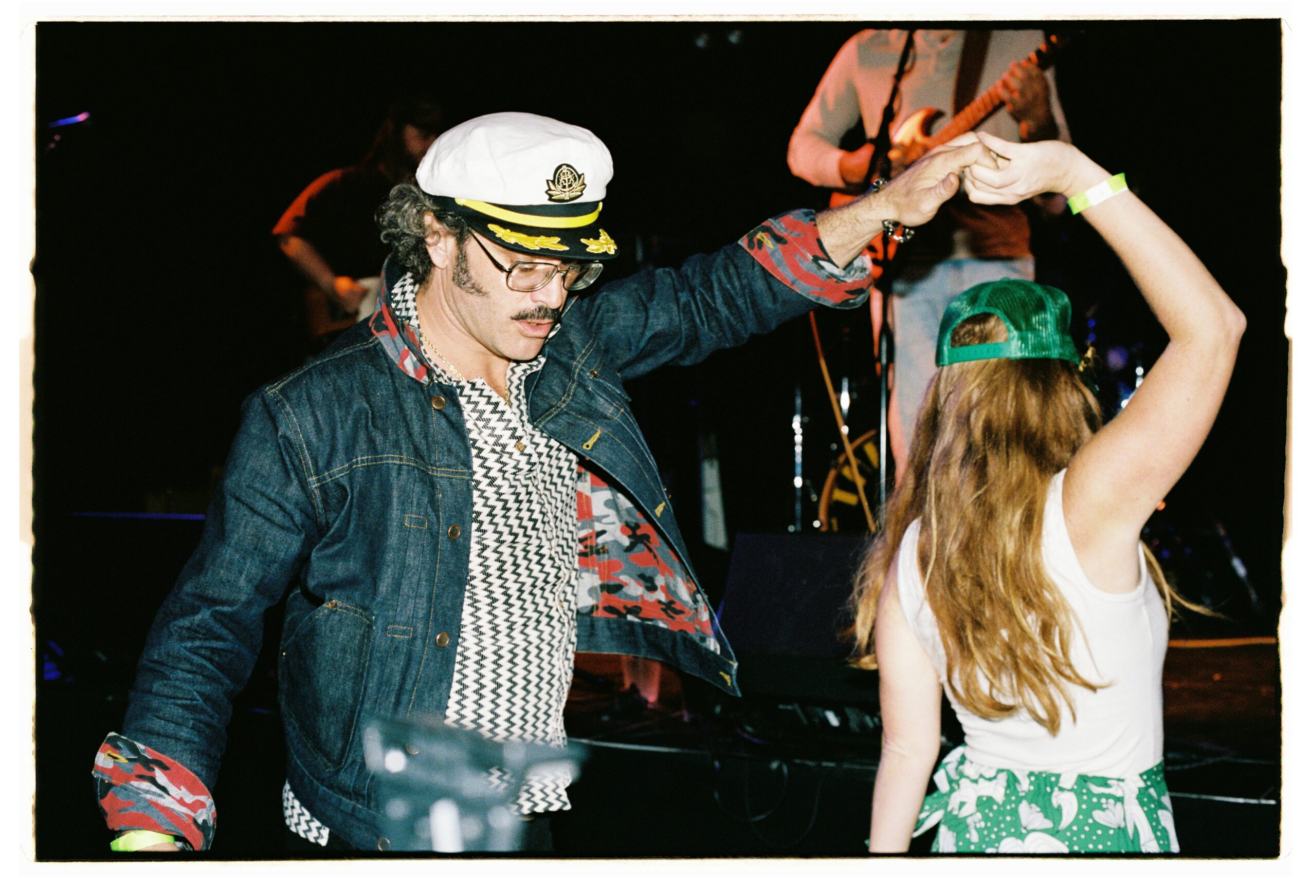 Person wearing a captain's hat dancing with a girl in a green cap, both raising their arms, dark background.