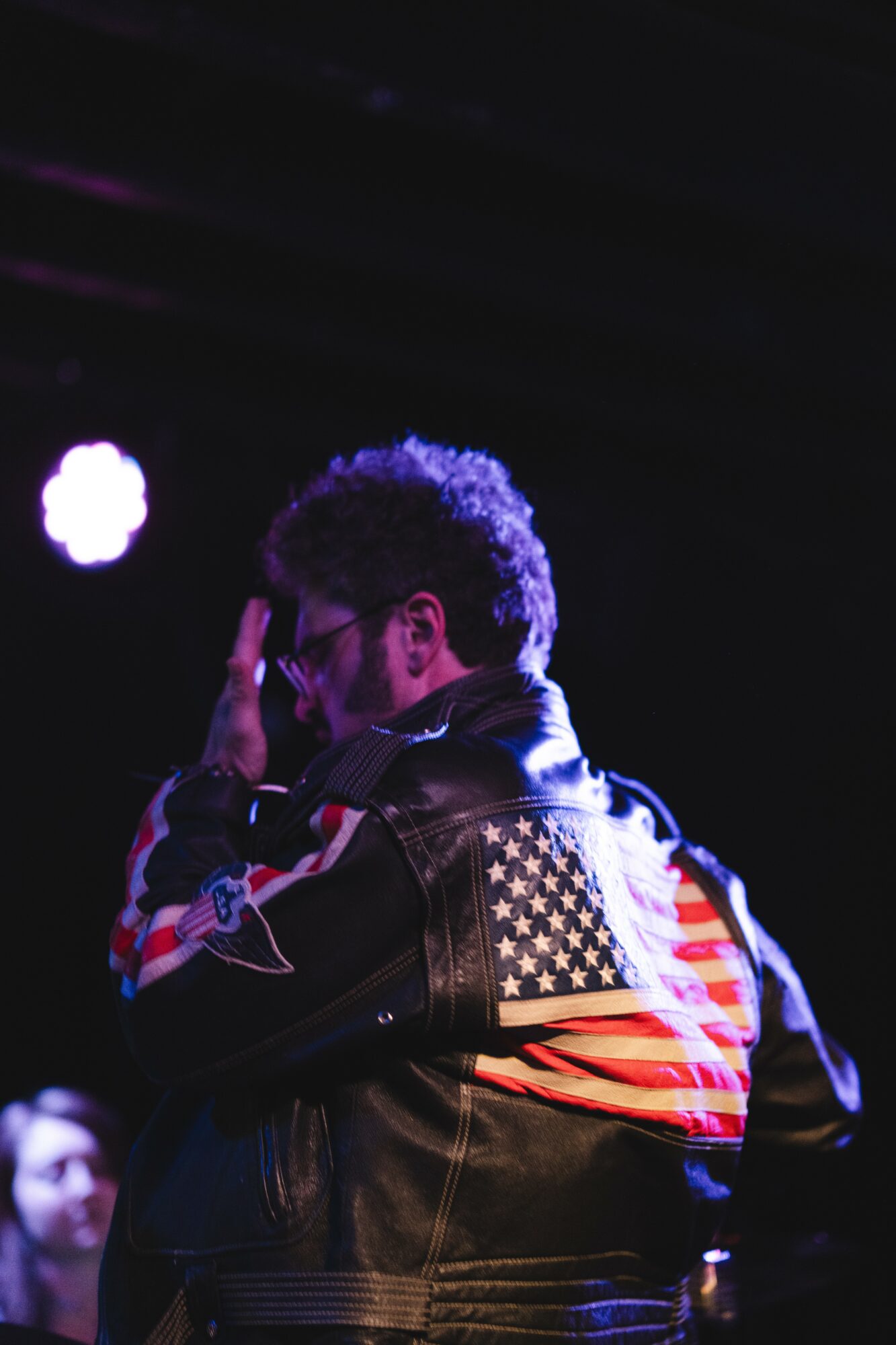 Person with curly hair and glasses wearing a jacket with American flag design, facing sideways, in a dark setting with stage lighting.