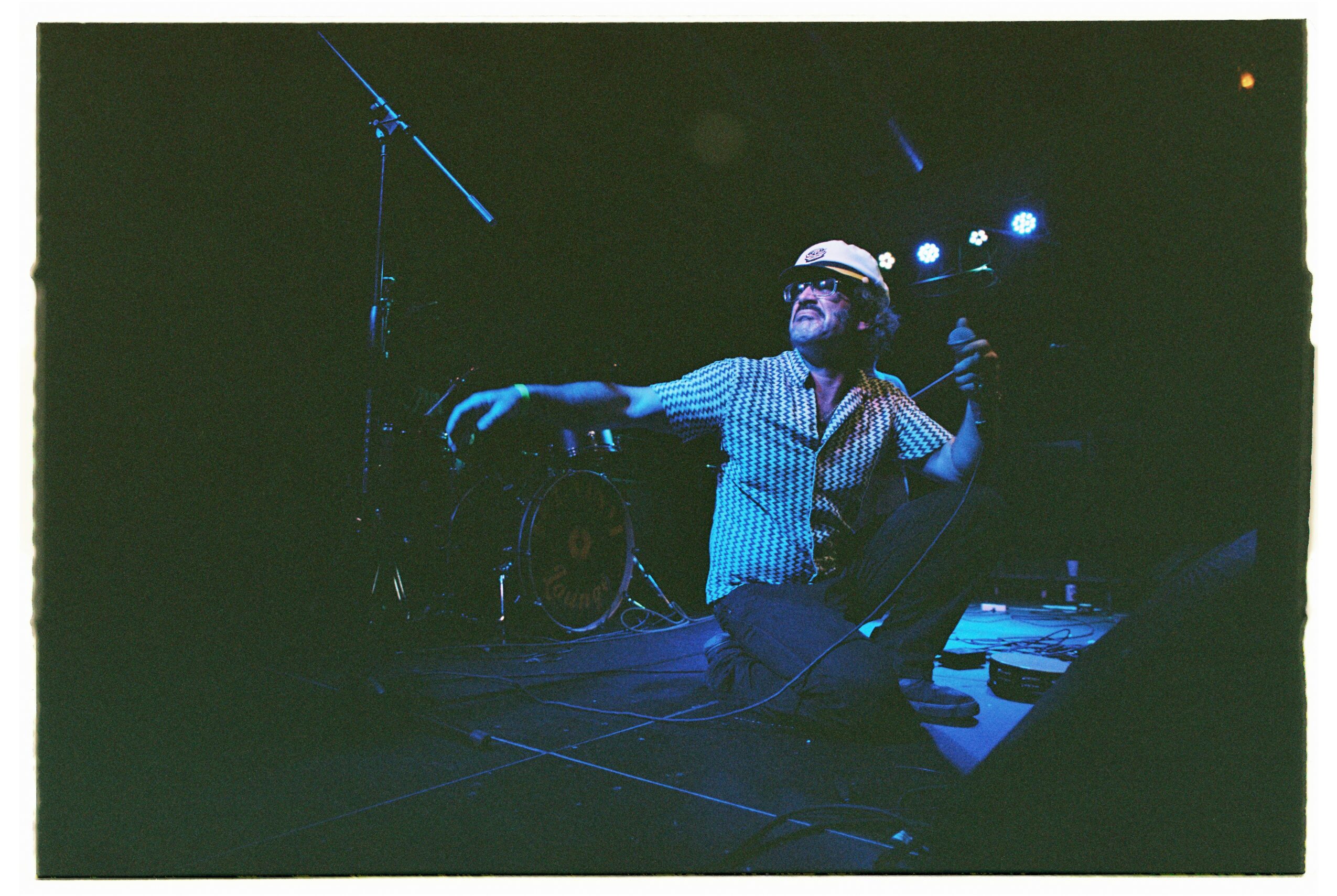 Person sitting on stage with musical equipment, wearing headphones, reaching out with one arm, dark background with stage lights.