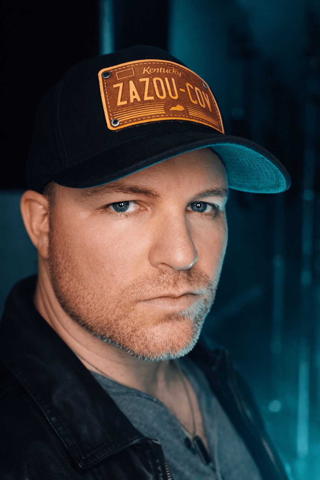 Close-up of a man wearing a black cap with a patch, looking serious, with a dark background.