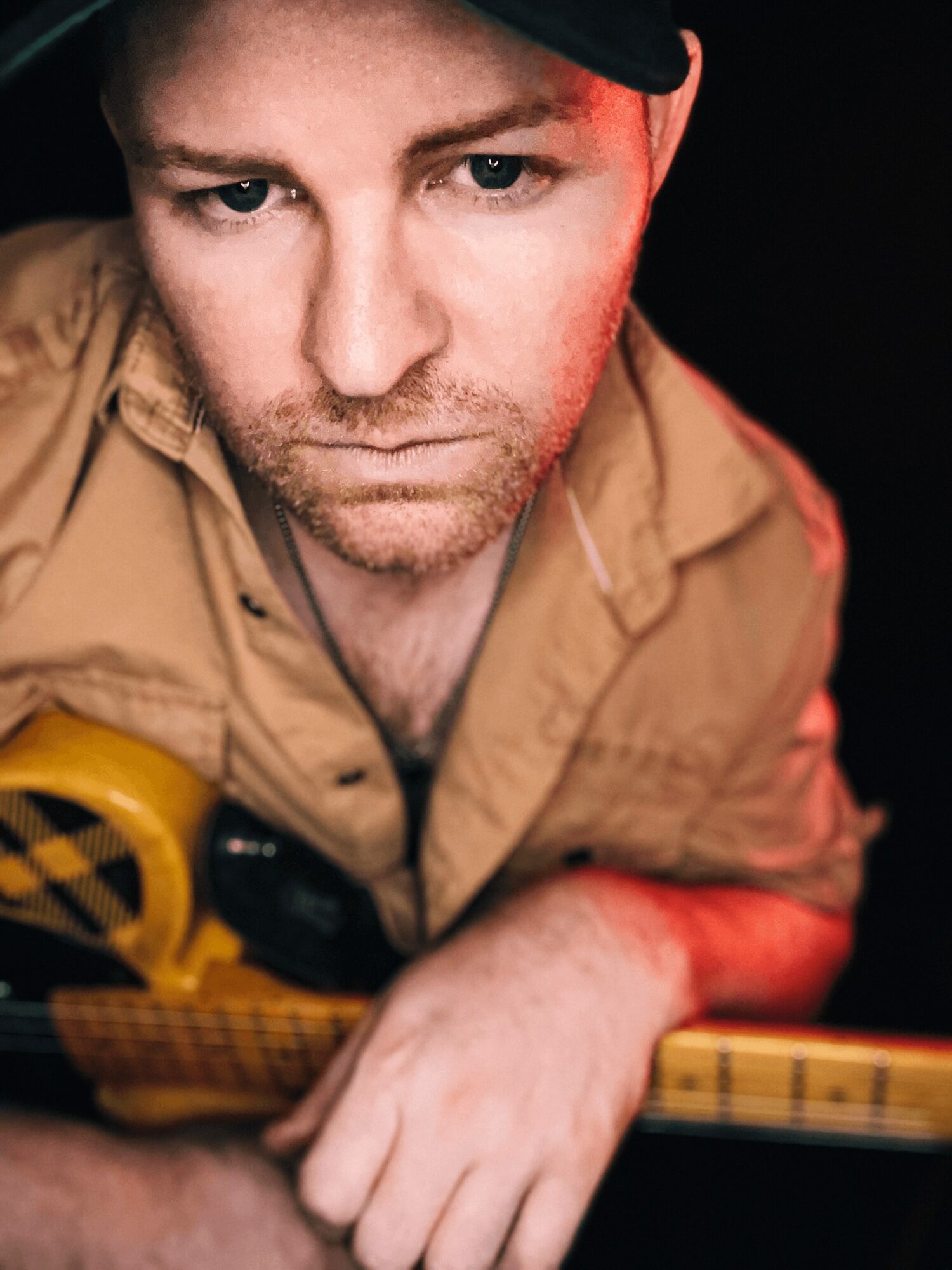Close-up of a man with a serious expression, wearing a tan shirt and a black cap, holding a guitar.