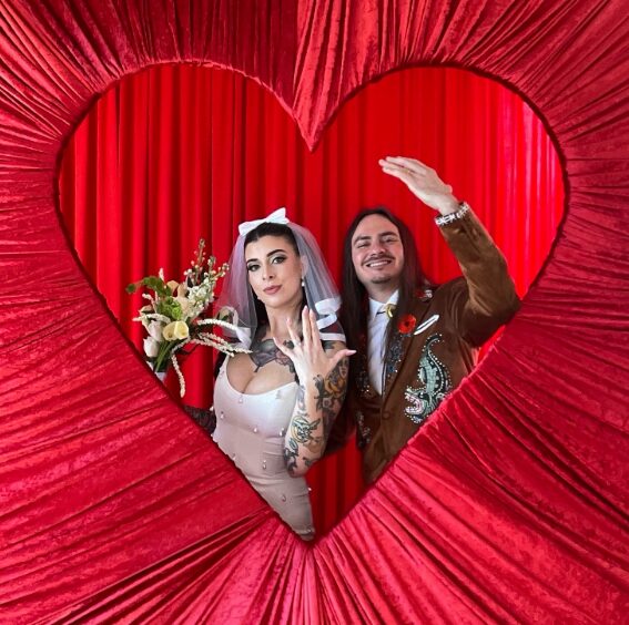 A bride and groom pose behind a large red heart-shaped frame with a red curtain background, smiling and waving.
