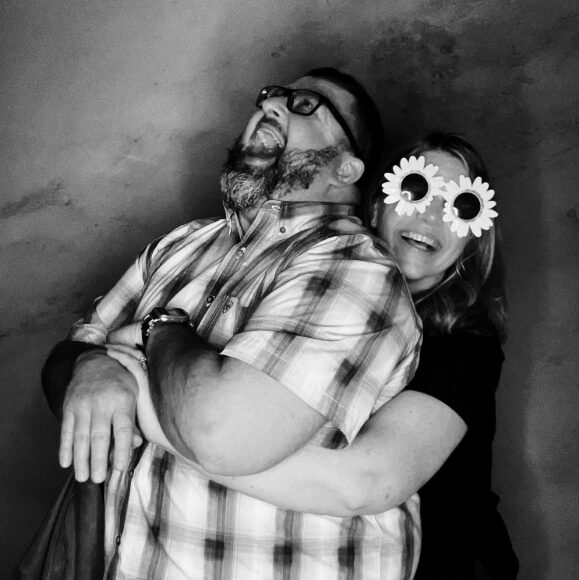 Two people laughing and hugging, one wearing glasses and a plaid shirt, the other with flower-shaped glasses, in black and white.