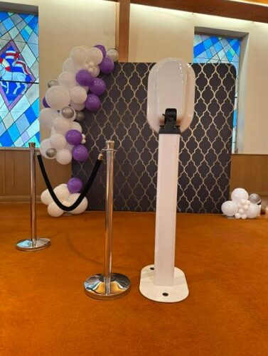 Decorative backdrop with purple and white balloons, a black and white patterned panel, and a white stand in front, inside a room with stained glass windows.