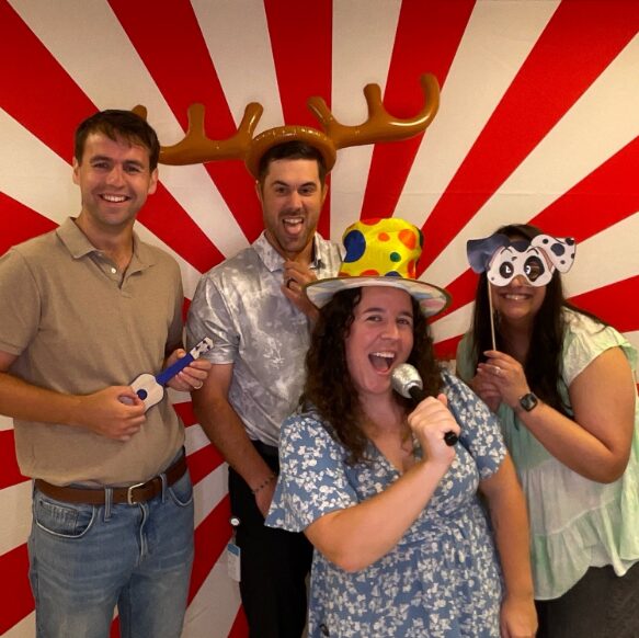 Four people smiling, one wearing a hat with colorful polka dots, another with a reindeer antler headband, holding props, in front of a red and white striped background.