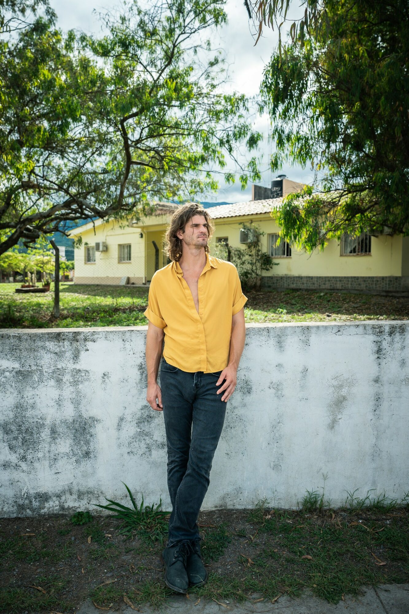 Young man in yellow shirt and jeans standing outdoors near a white wall, trees and a building in background.