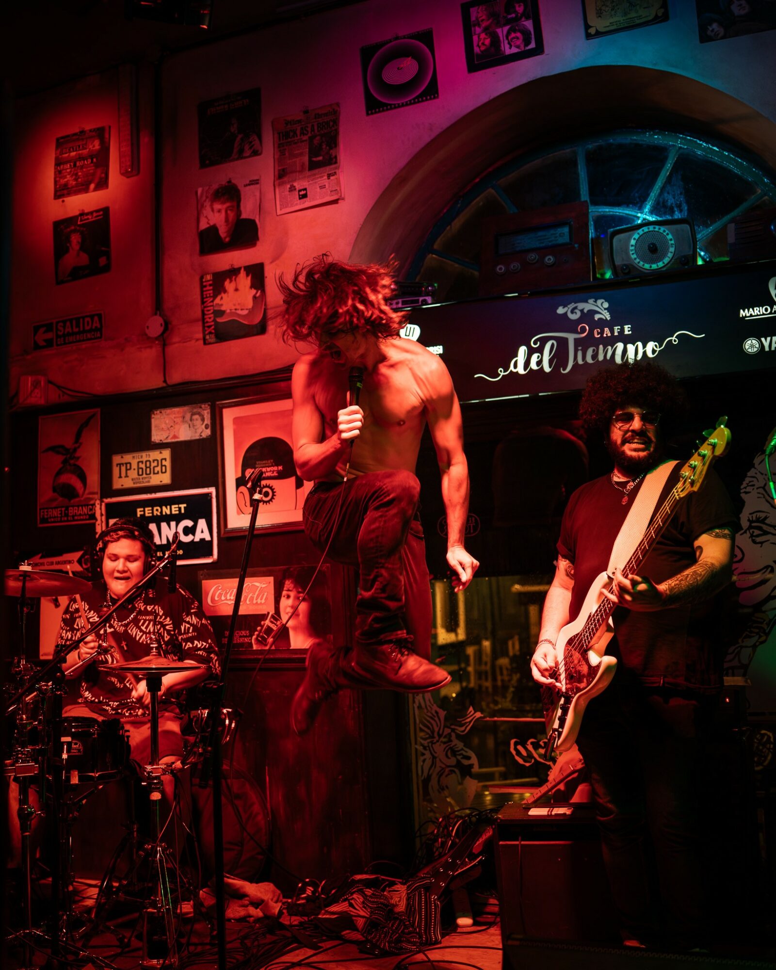 Three musicians perform on stage with colorful lighting, surrounded by framed pictures and a neon sign, in a cozy venue.