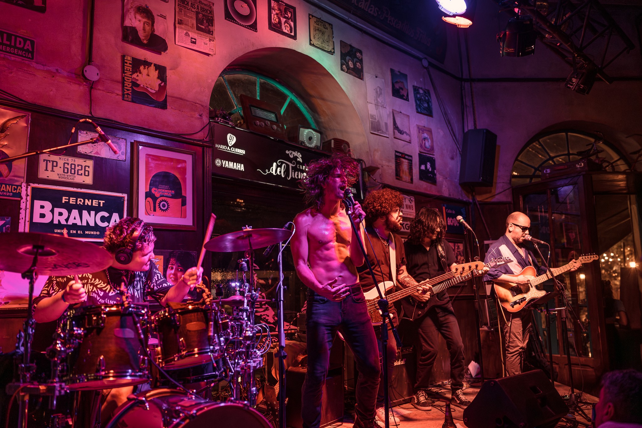 Band performs live on stage with instruments, colorful lighting, and framed pictures on the wall behind them.