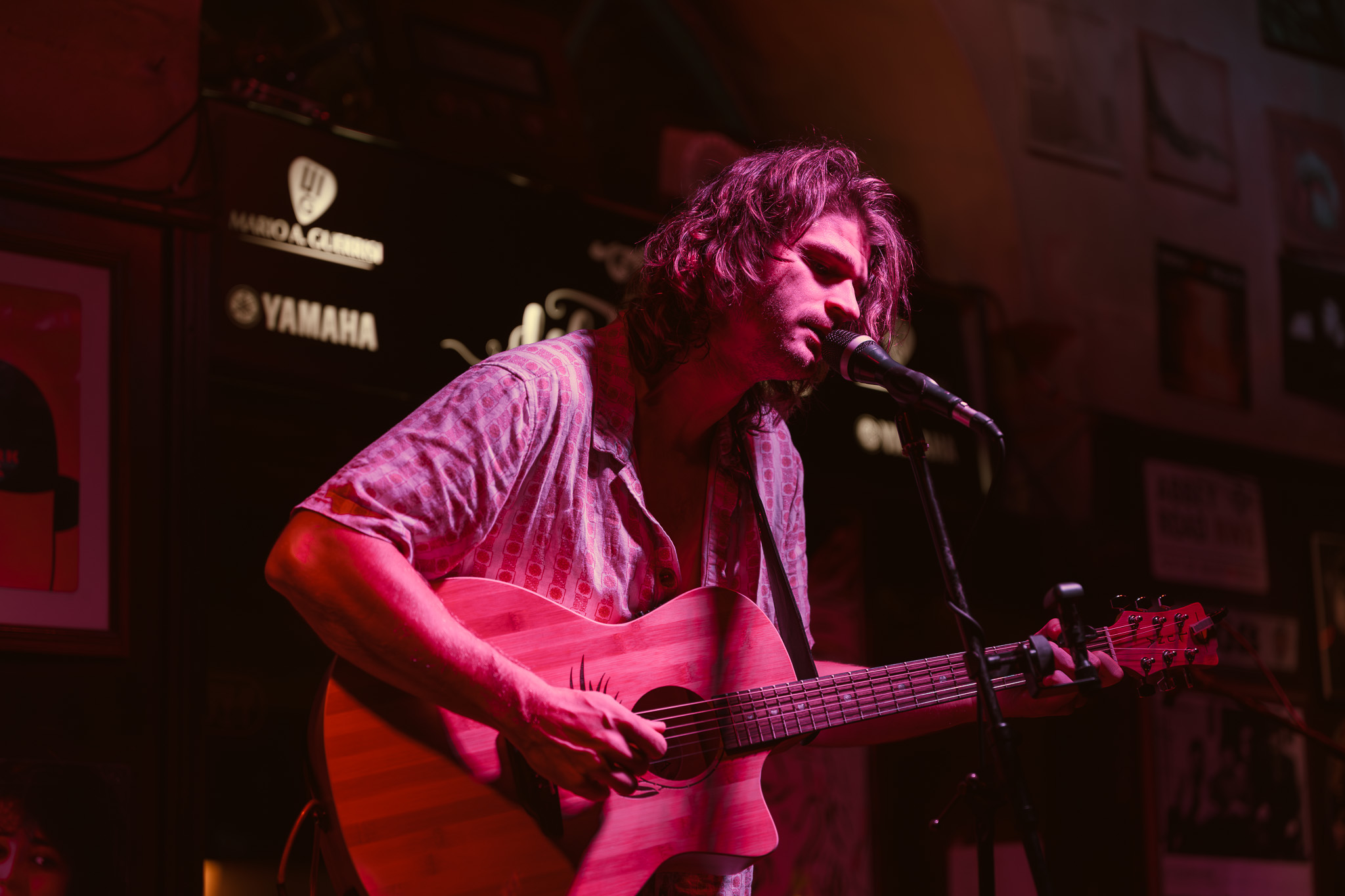 Person with curly hair playing guitar and singing into a microphone on stage, dimly lit with posters on the wall behind.