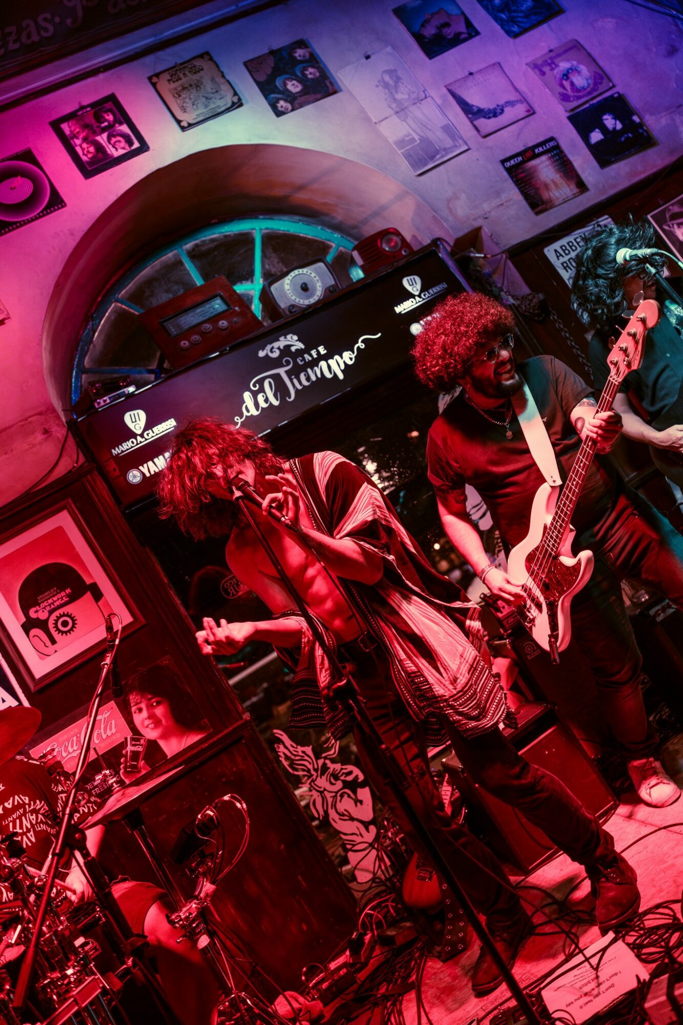 Two musicians perform on stage with a keyboard and guitar, surrounded by posters and equipment, in a colorful venue.