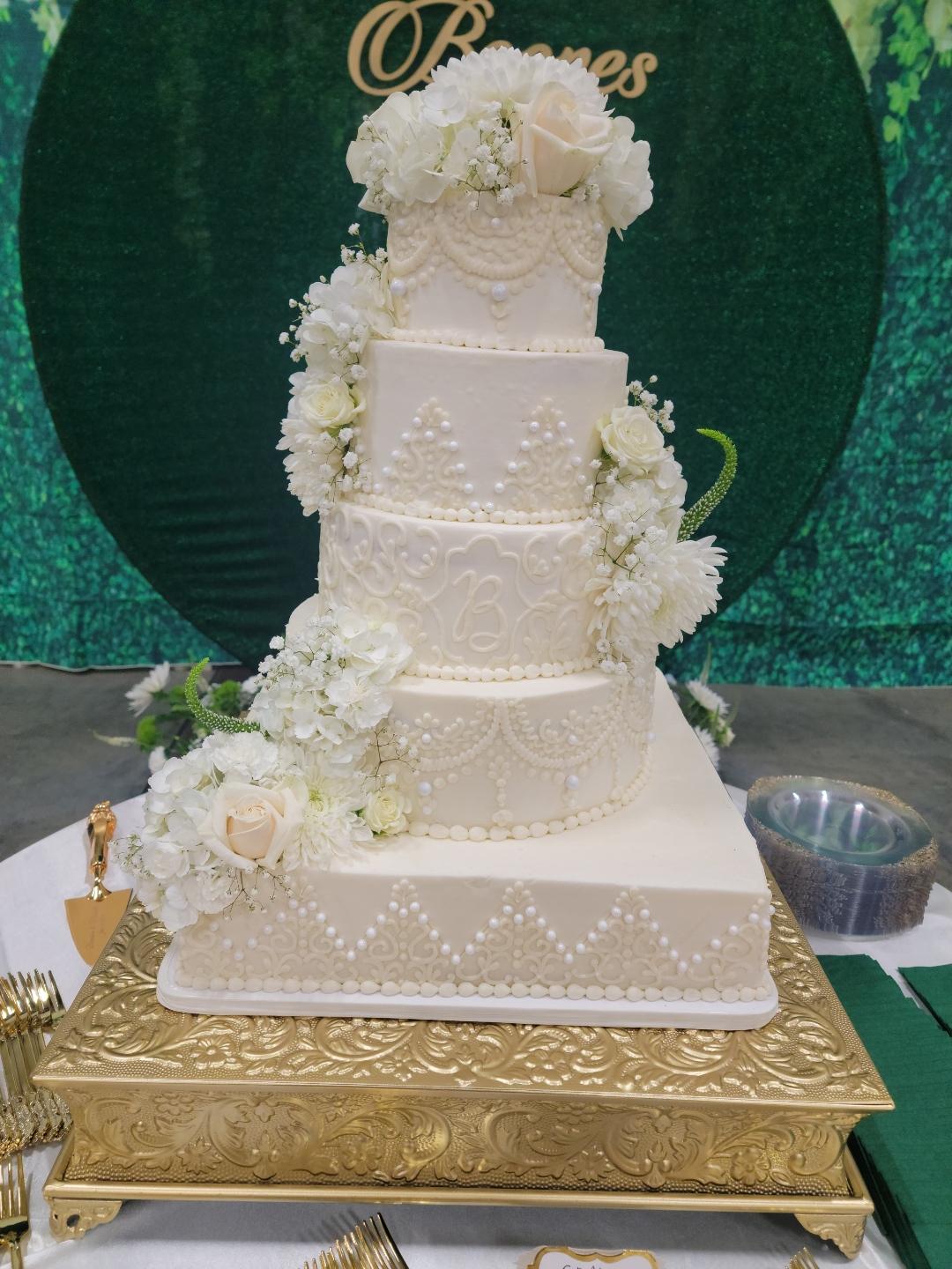 Four-tier white wedding cake decorated with white flowers, on a gold stand, with a green backdrop.