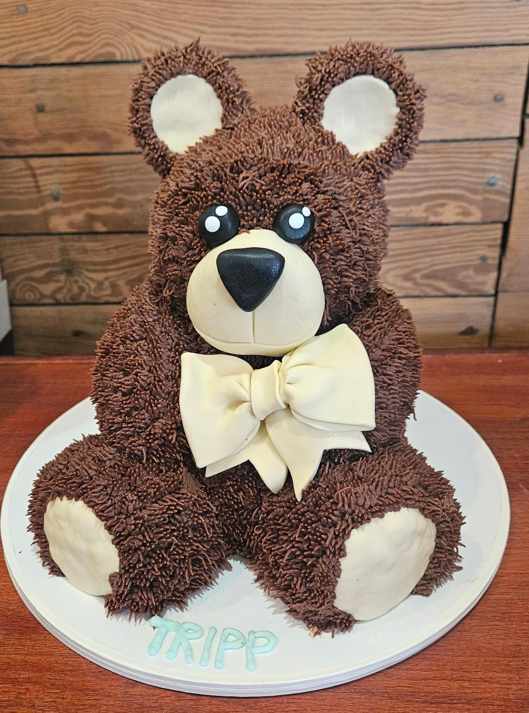 Bear-shaped cake with a cream-colored bow tie, sitting on a white plate with the name 'TRIPP' written on it.