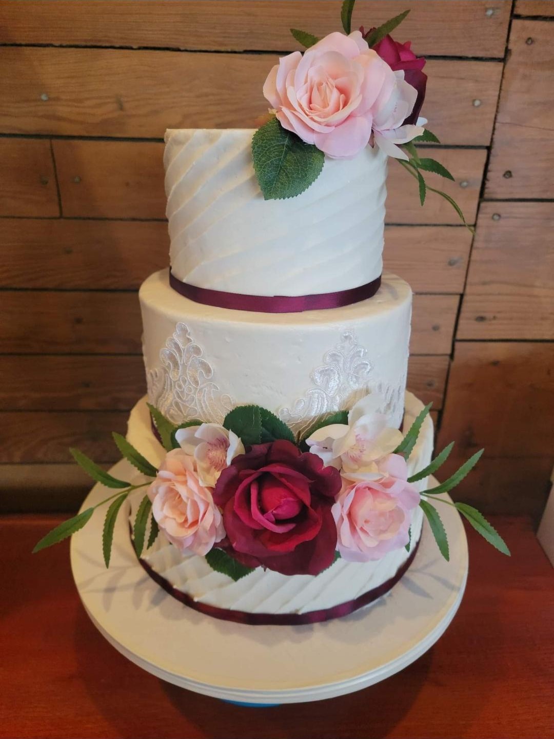Two-tiered cake with pink and burgundy flowers, green leaves, on a white plate against a wooden background.