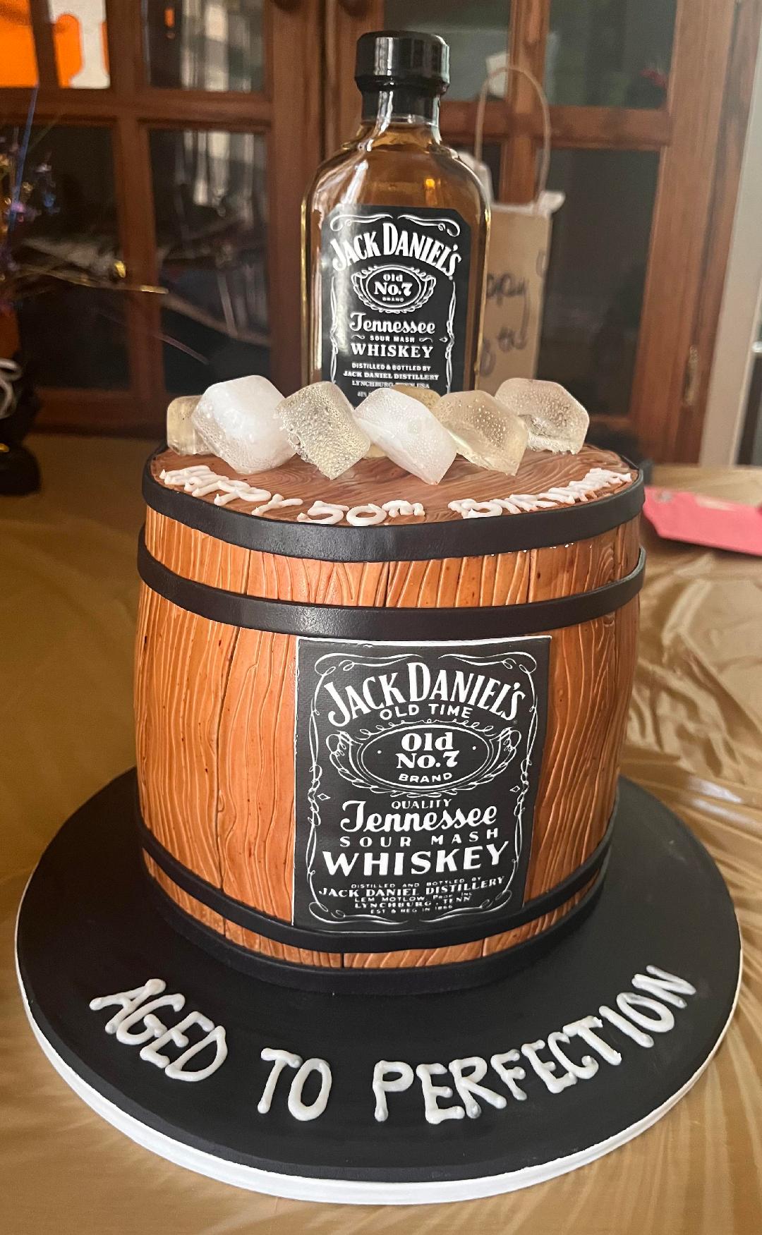 Cake decorated as a whiskey barrel with a Jack Daniel's bottle on top, surrounded by sugar cubes, on a black base with white text.