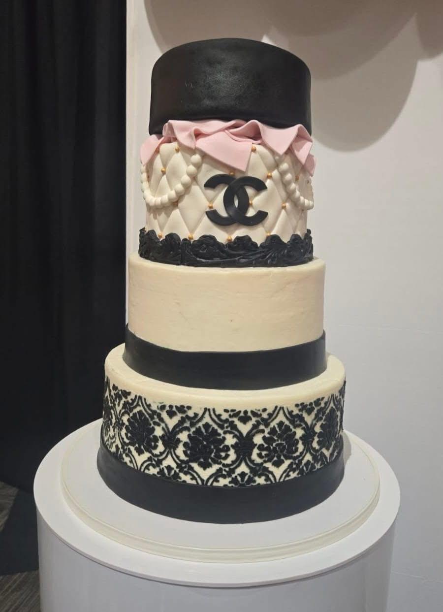 Four-tiered cake with black, white, and pink decorations, including a pink bow, pearls, and a Chanel logo, on a white stand.