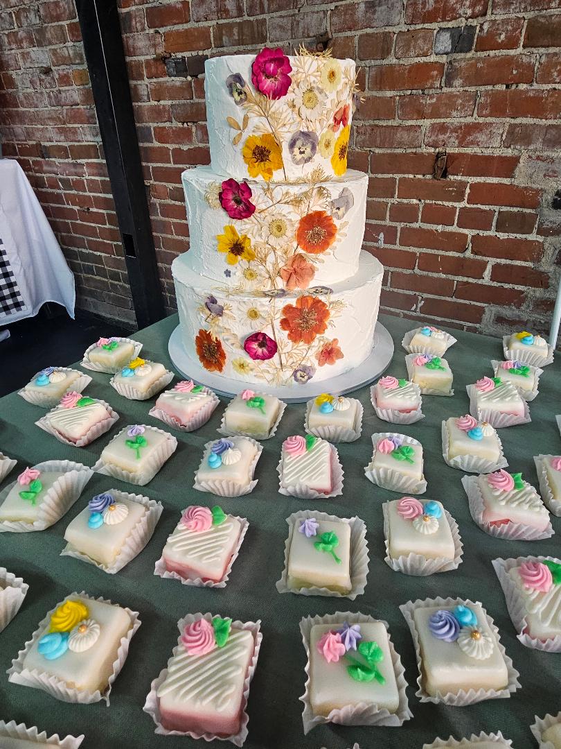 Three-tiered cake decorated with colorful flowers, surrounded by decorated cookies on a table against a brick wall.