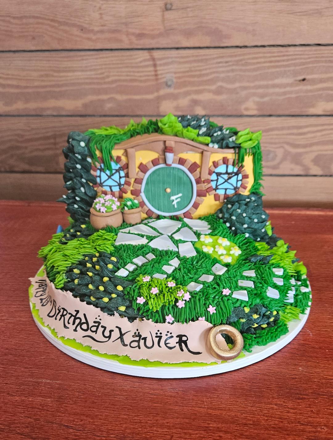 Decorative birthday cake with a garden theme, featuring a door, pathway, and greenery, on a wooden surface. Text reads 'Happy Birthday Xavier'.
