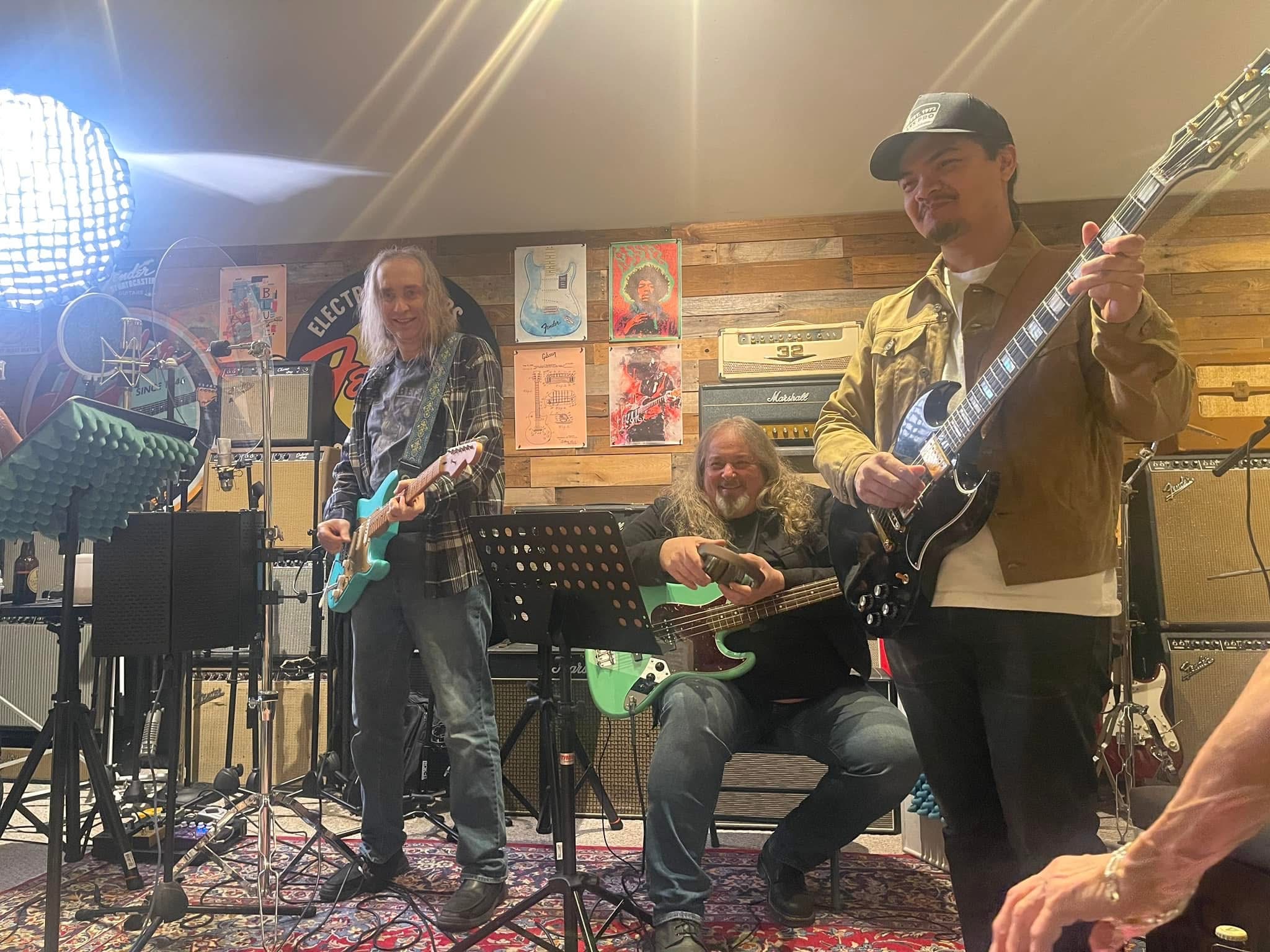 Three musicians perform on stage with guitars, a keyboard, and a microphone, in a cozy indoor setting with artwork on the wall.