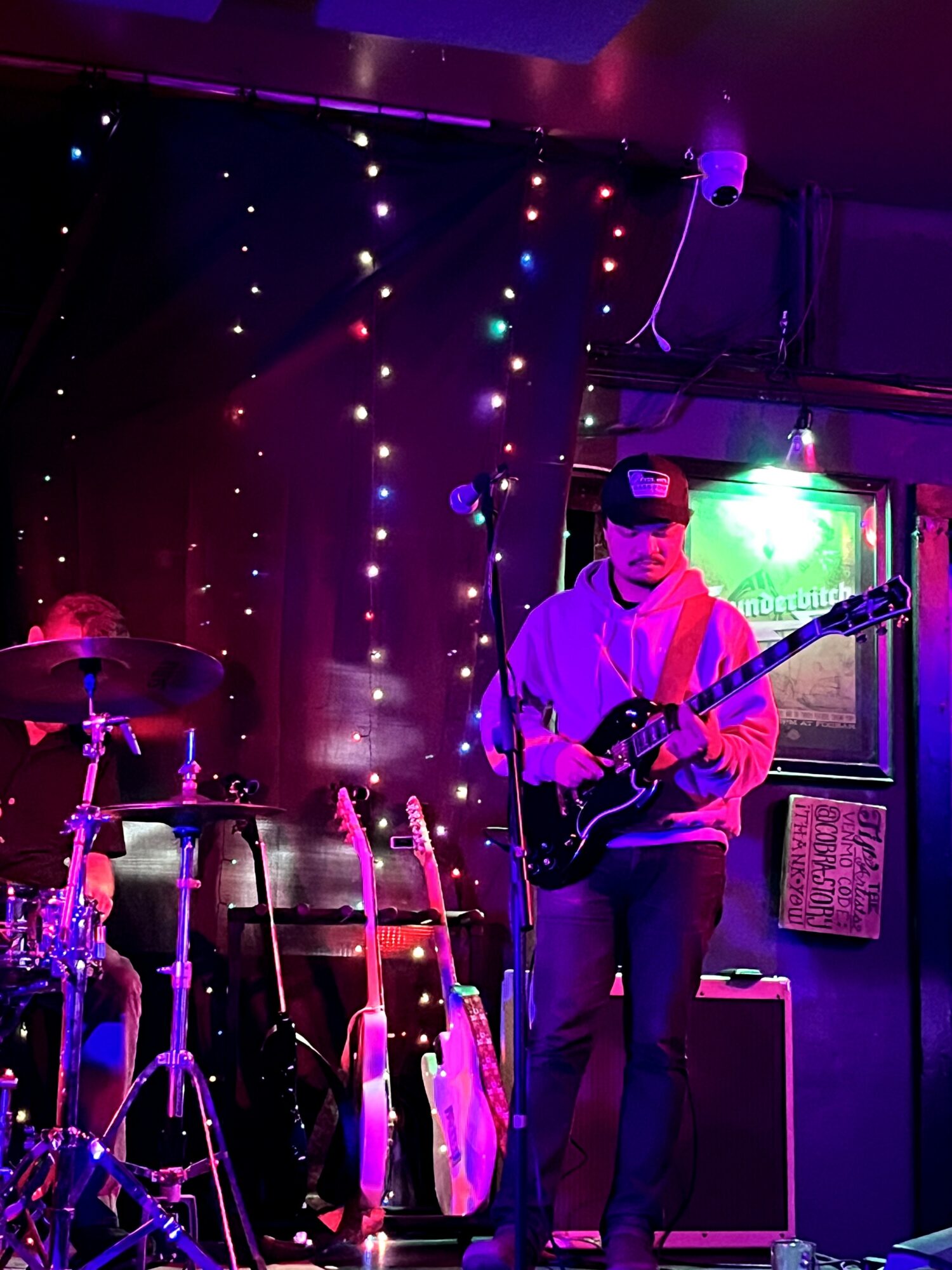 Musician playing electric guitar on stage with colorful lights and musical instruments in background.
