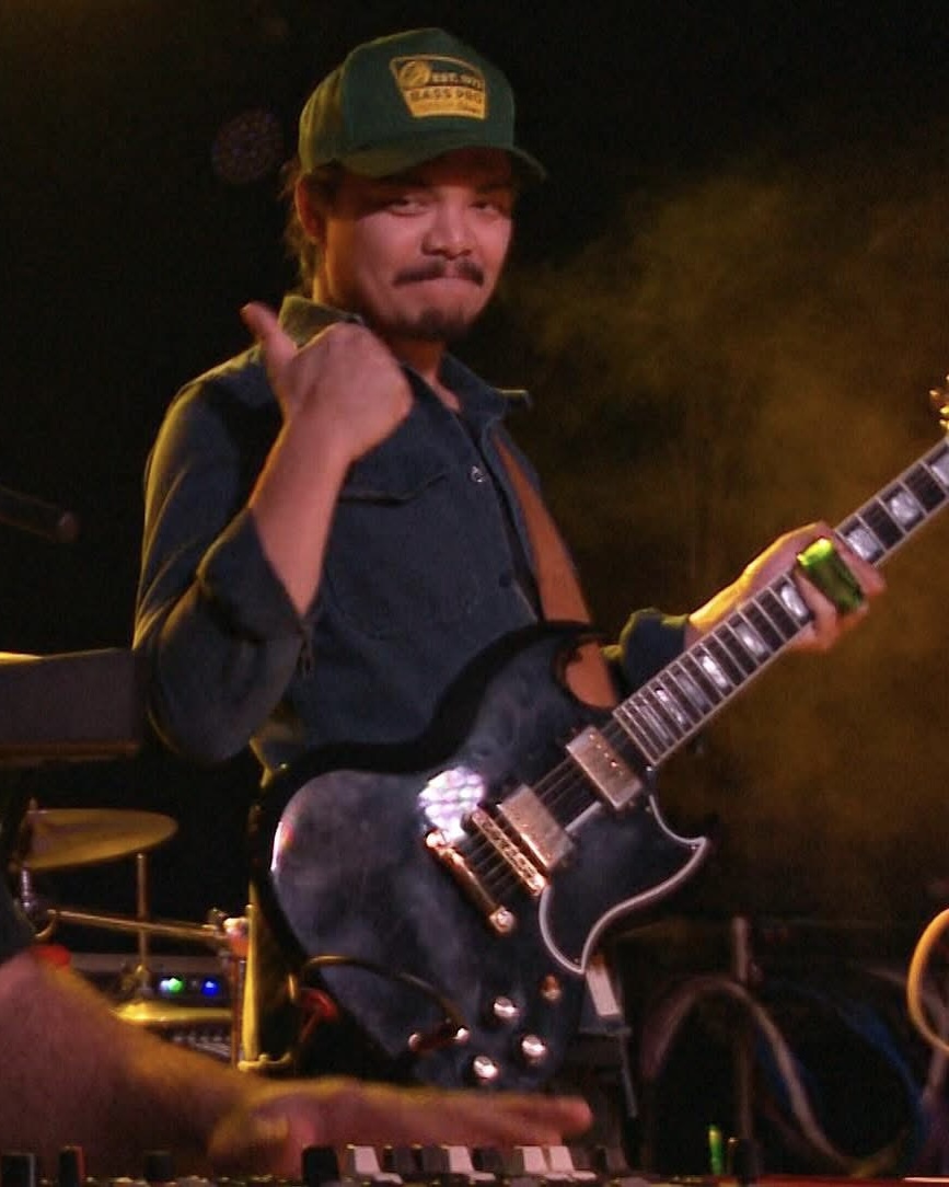 Person with a beard and mustache wearing a green cap and denim shirt, holding a black electric guitar, on stage with warm lighting.