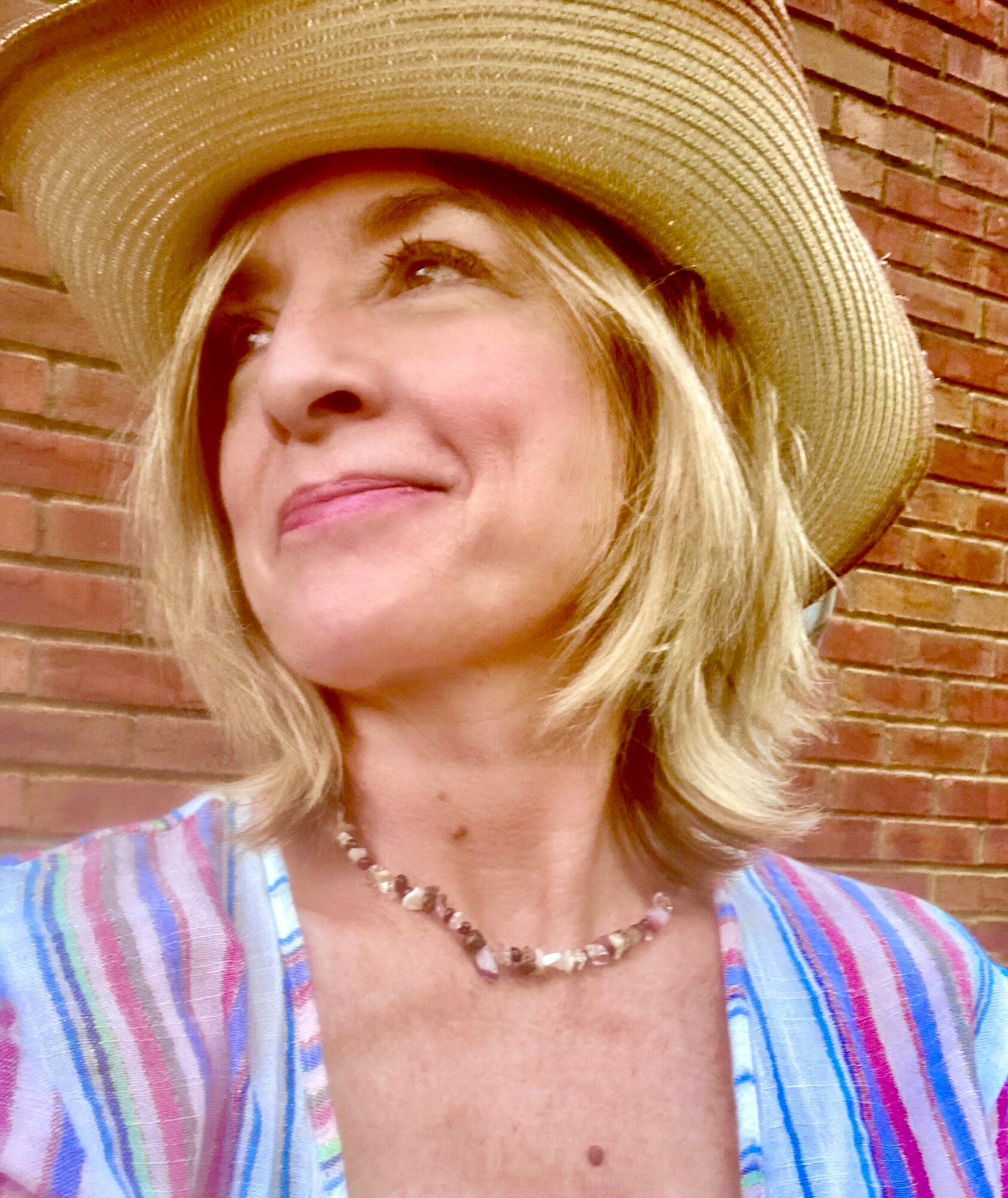 Woman wearing a wide-brimmed hat and colorful striped shirt, smiling, with a brick wall background.