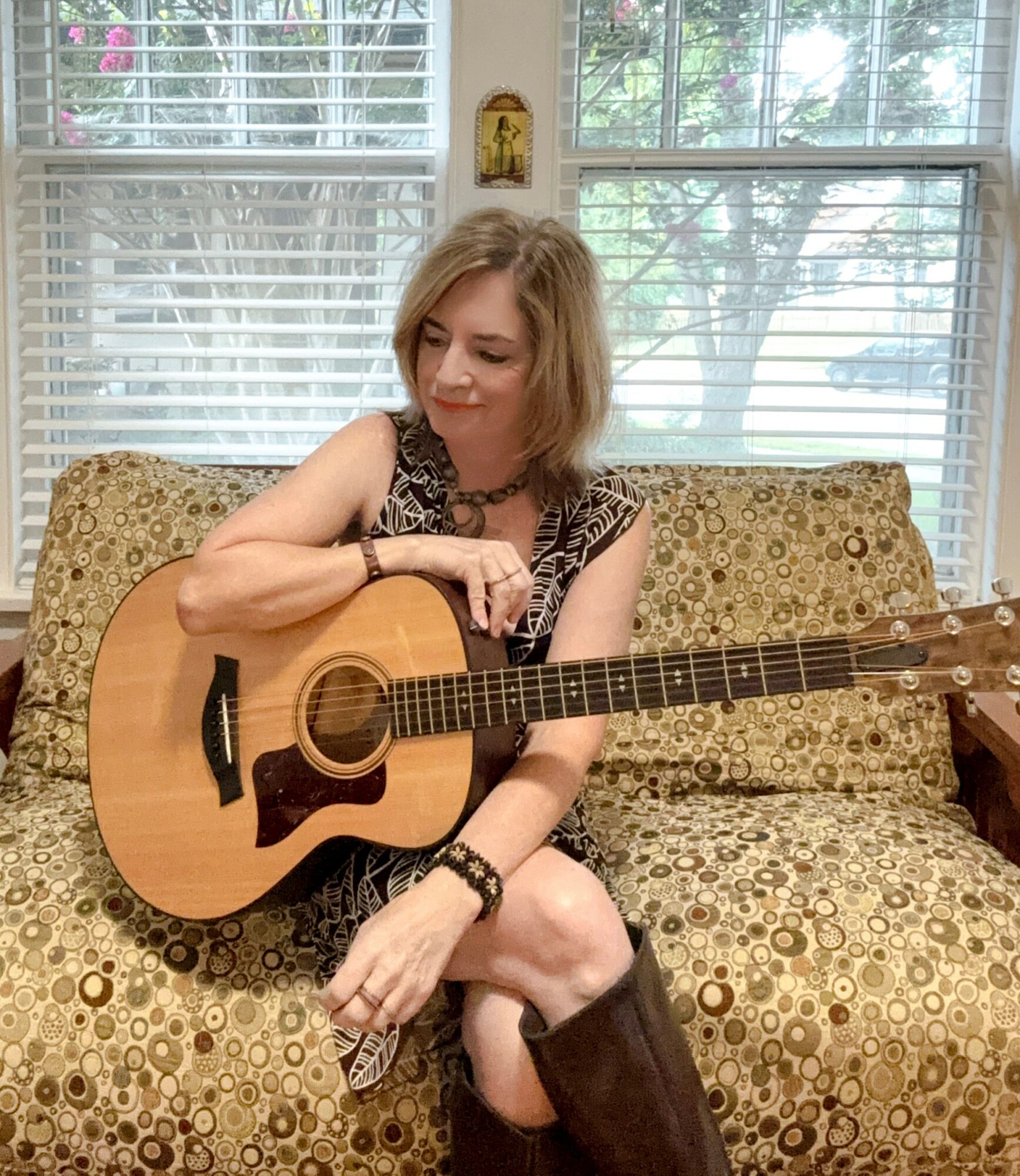 Woman sitting on a patterned couch holding an acoustic guitar, with windows and trees in the background.