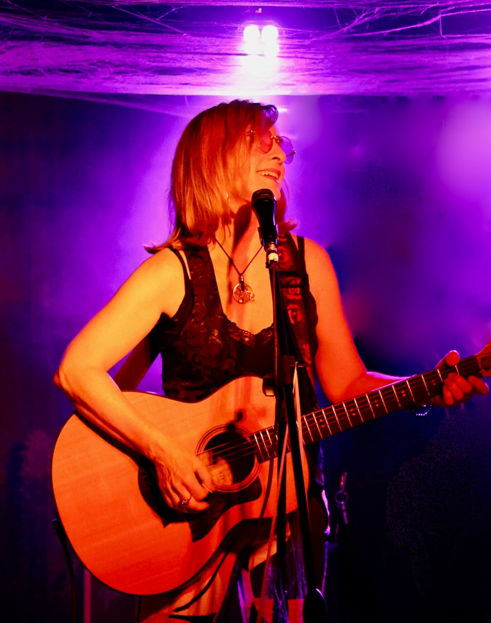 Woman with red hair singing into a microphone and playing an acoustic guitar on stage, purple lighting background.