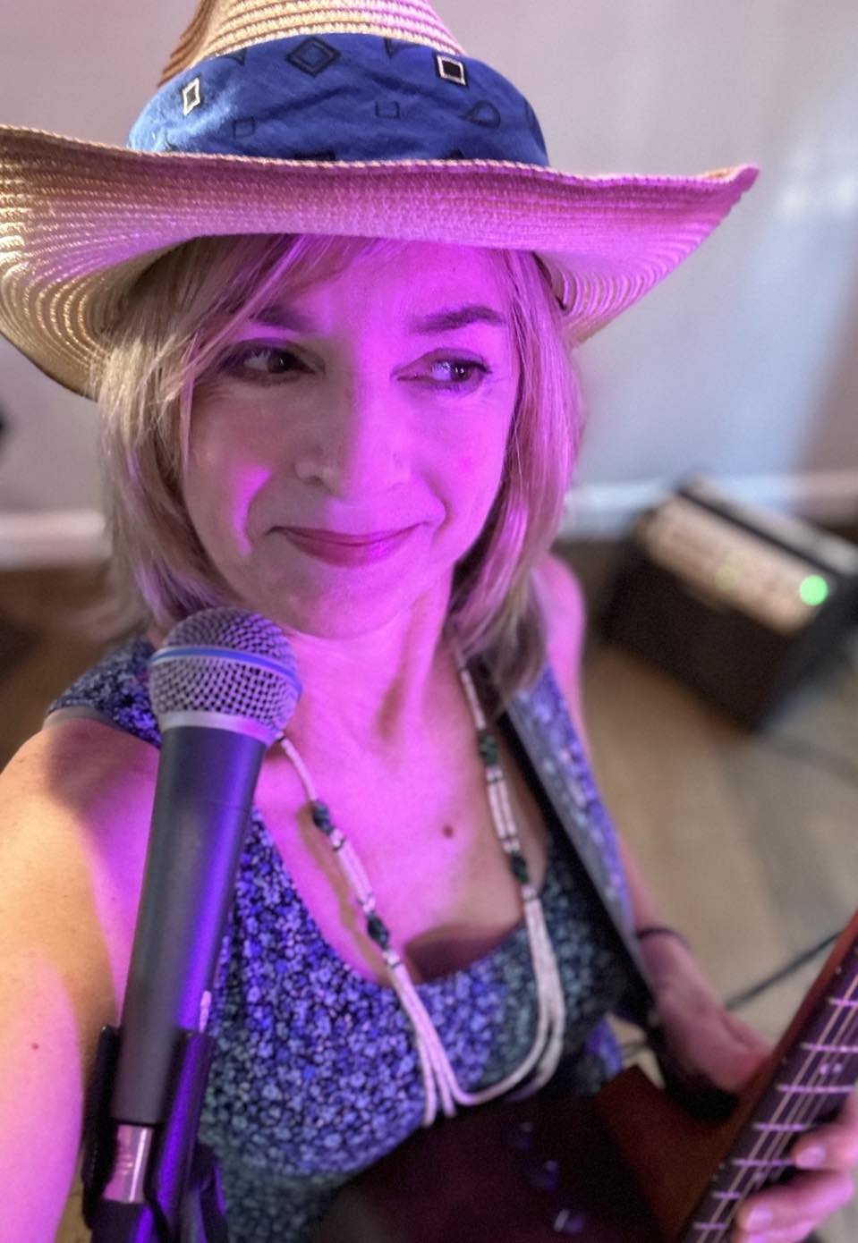 Woman wearing a large hat, smiling, holding a guitar, with a microphone nearby, in a room with a blurred background.