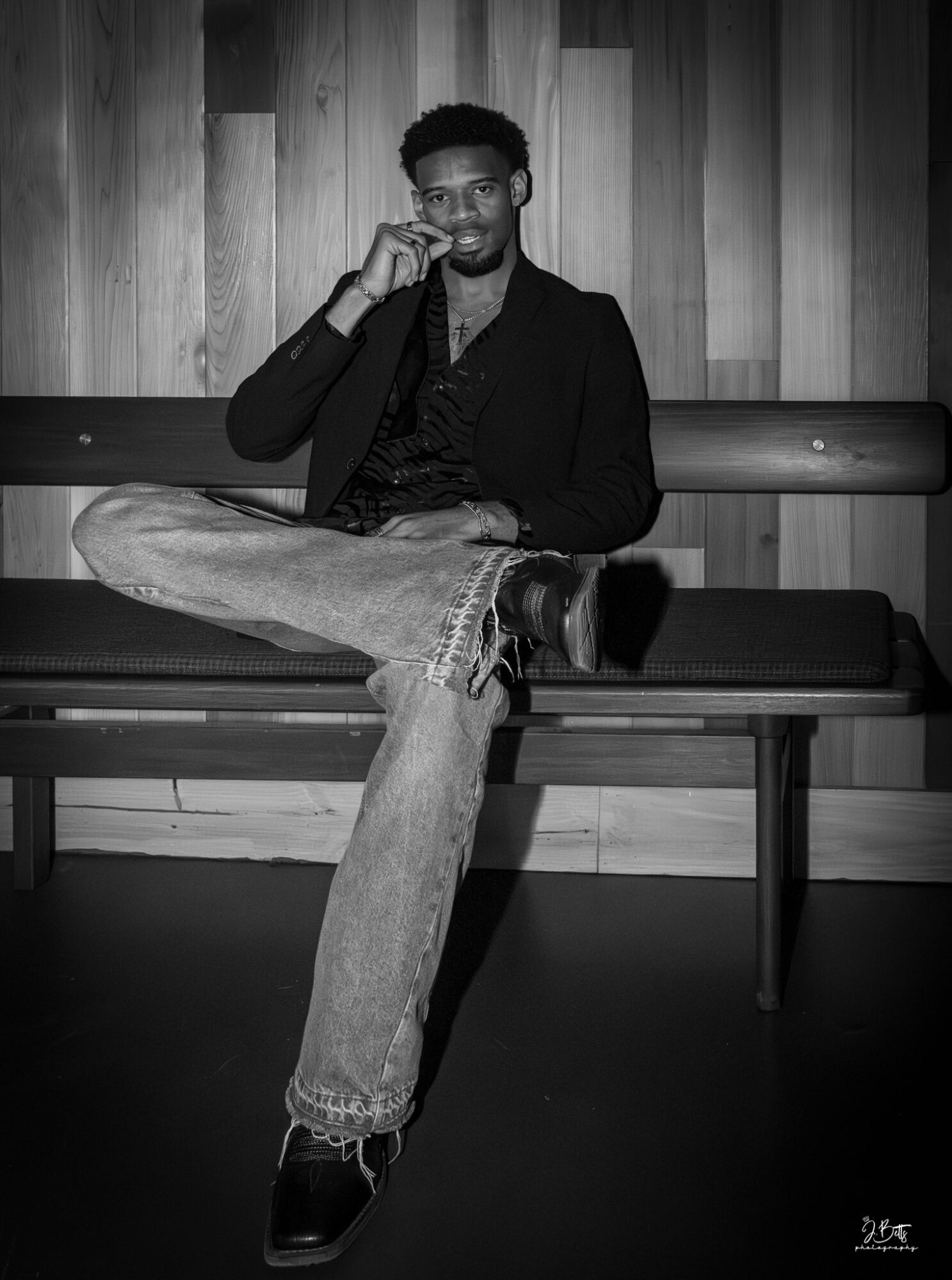 Young man sitting on a bench with one leg crossed over the other, smiling, in front of a wooden wall, black and white.