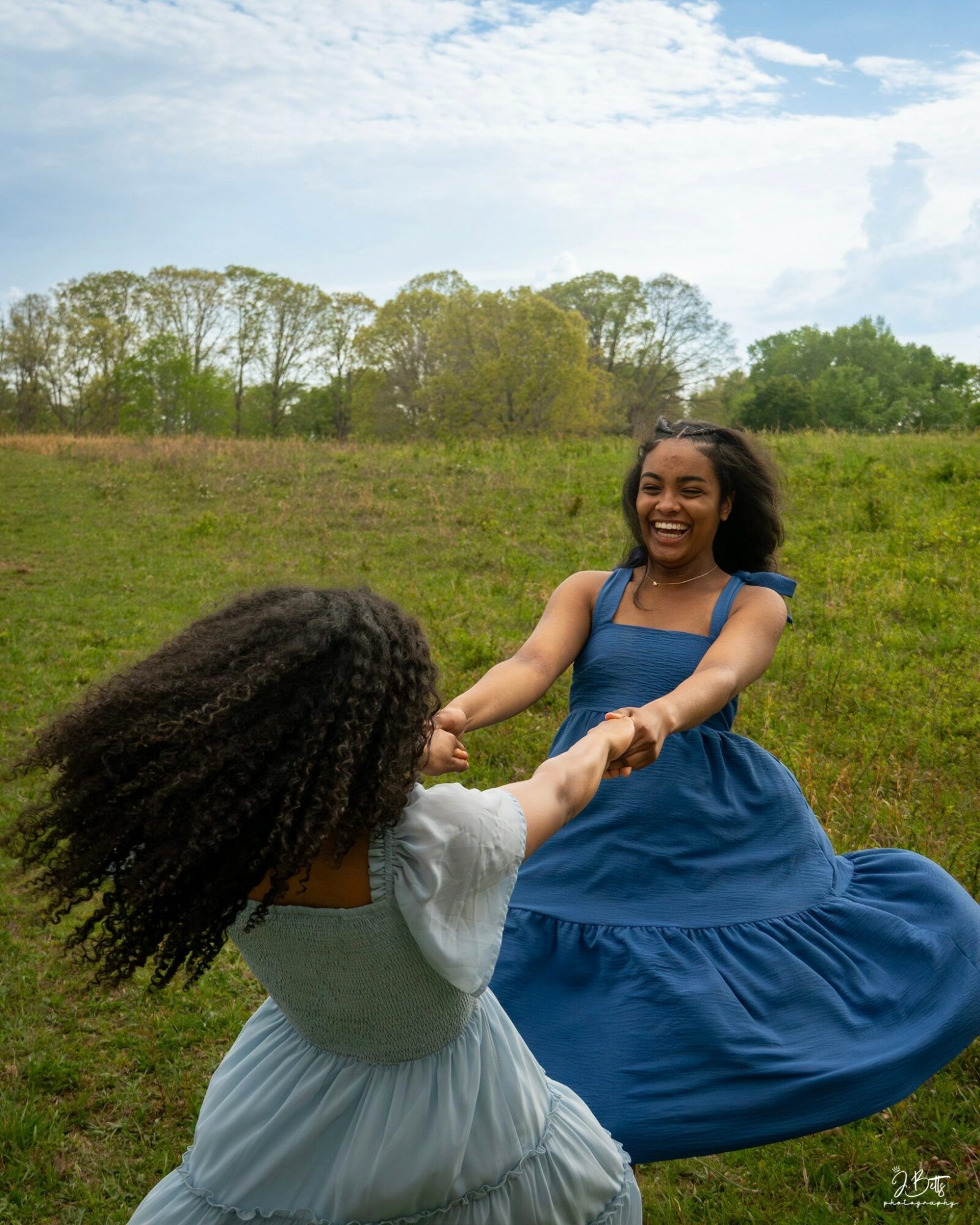 Two women holding hands and spinning outdoors in a grassy field with trees and blue sky.