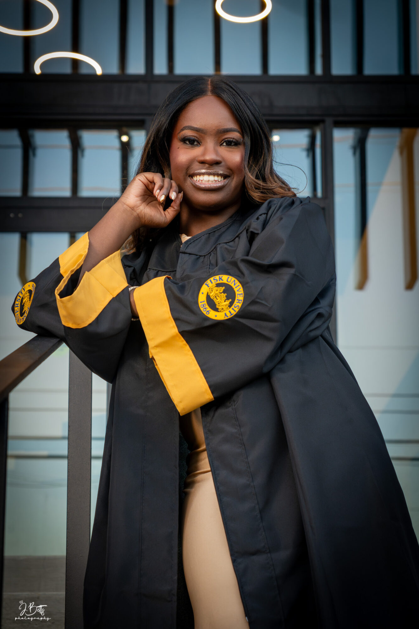 Young woman in graduation gown smiling outdoors, standing in front of a modern building with glass and metal elements.