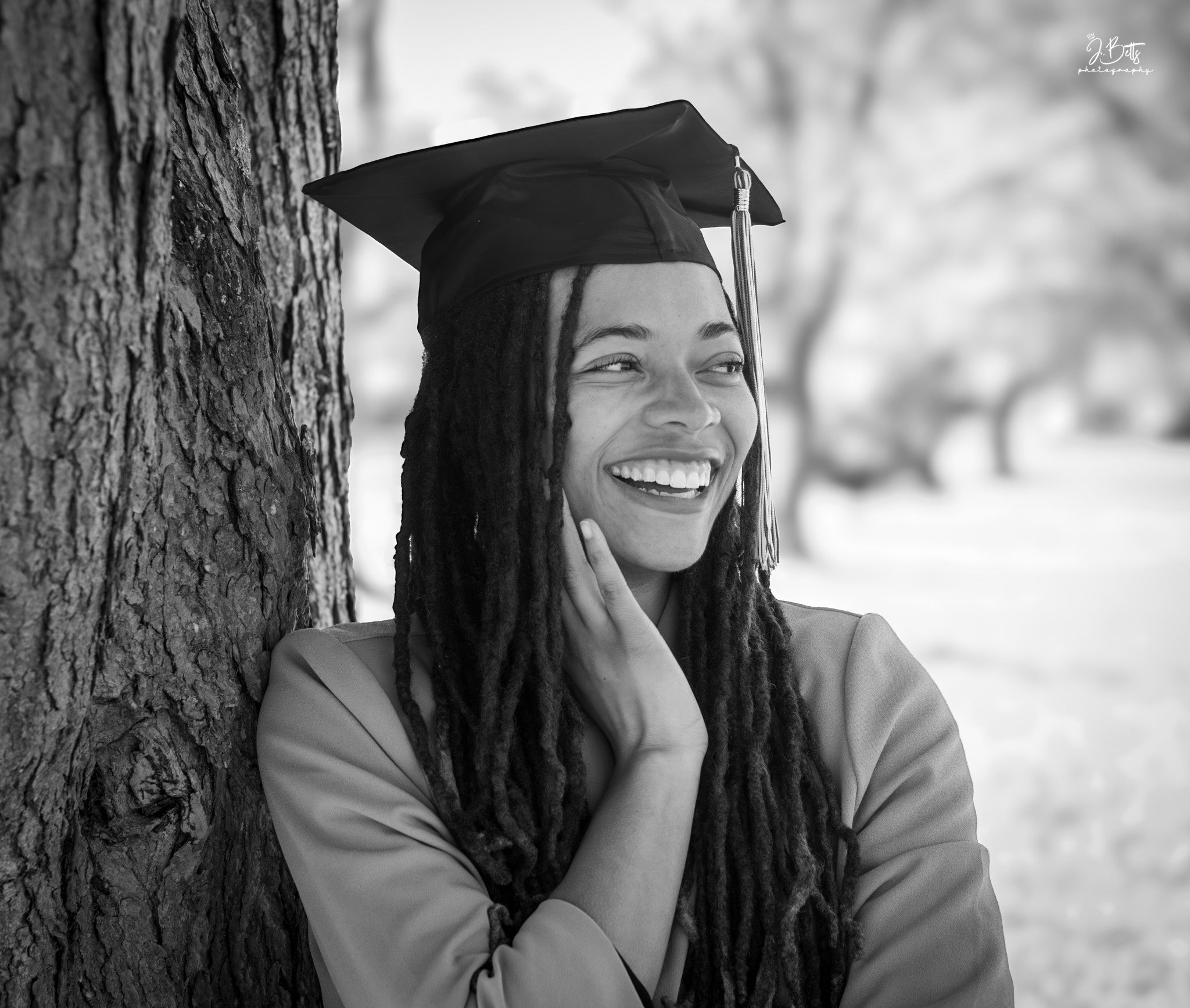 Young woman with dreadlocks wearing a graduation cap, smiling and touching her face outdoors near a tree.