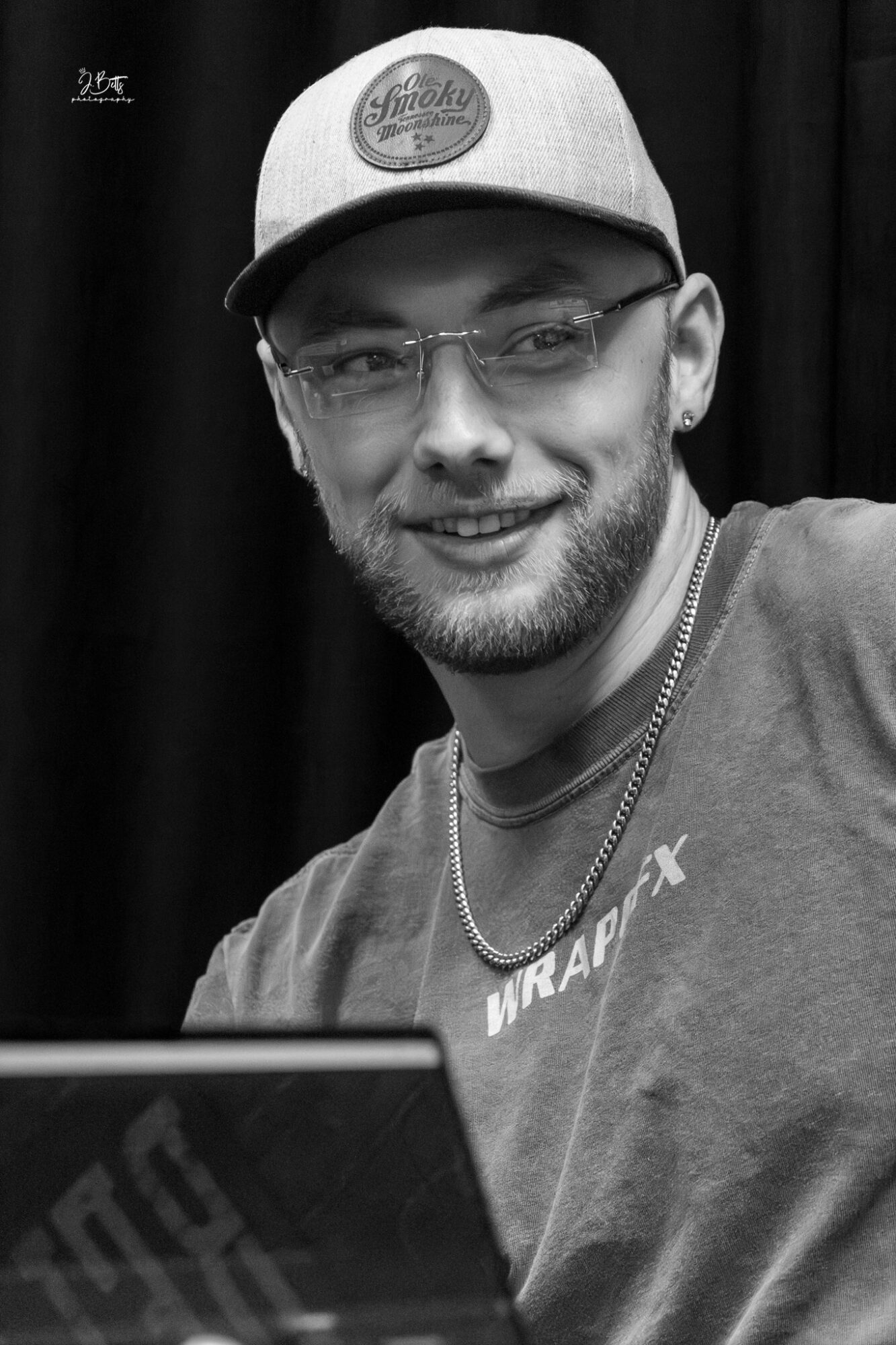 Young man with glasses and a cap smiling, wearing a T-shirt and chain, in front of a dark background.
