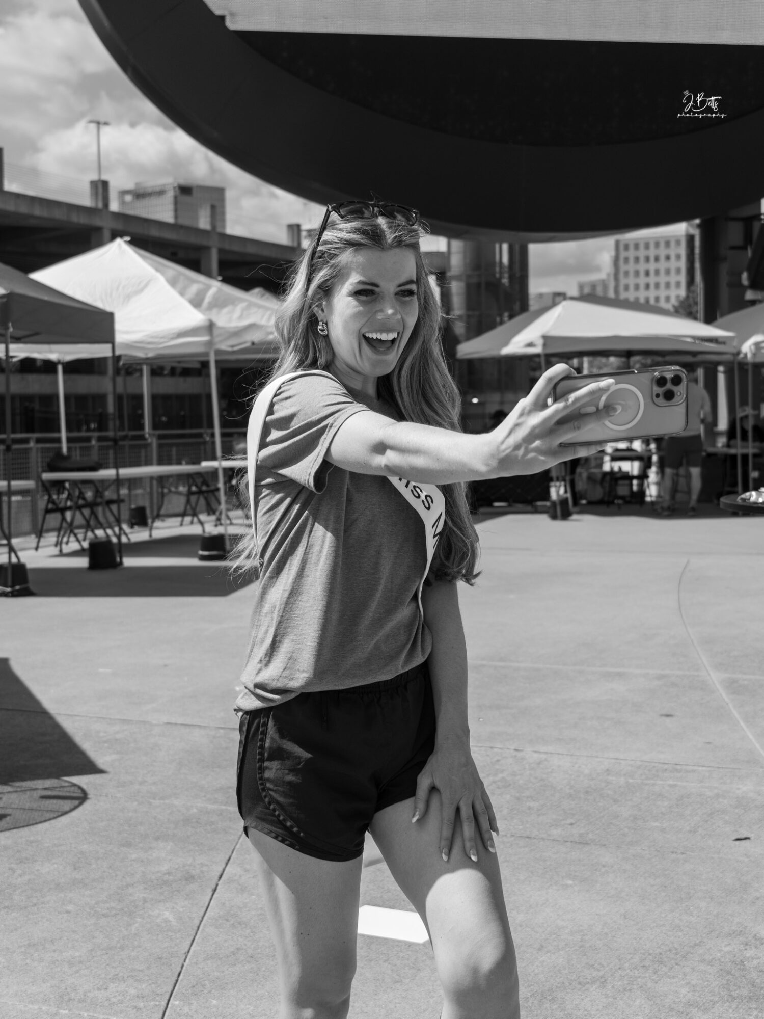 Young woman smiling and extending her arm outdoors with tents and buildings in background.