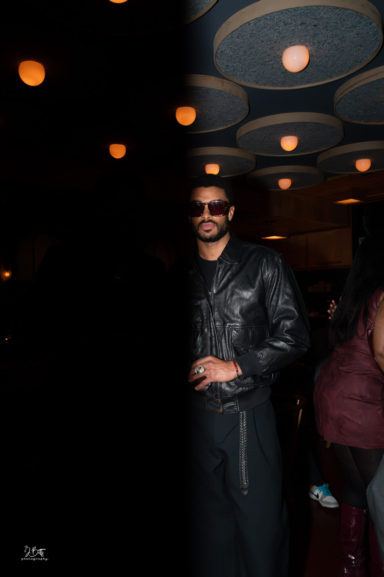 Man wearing sunglasses and a leather jacket standing indoors with ceiling lights above.