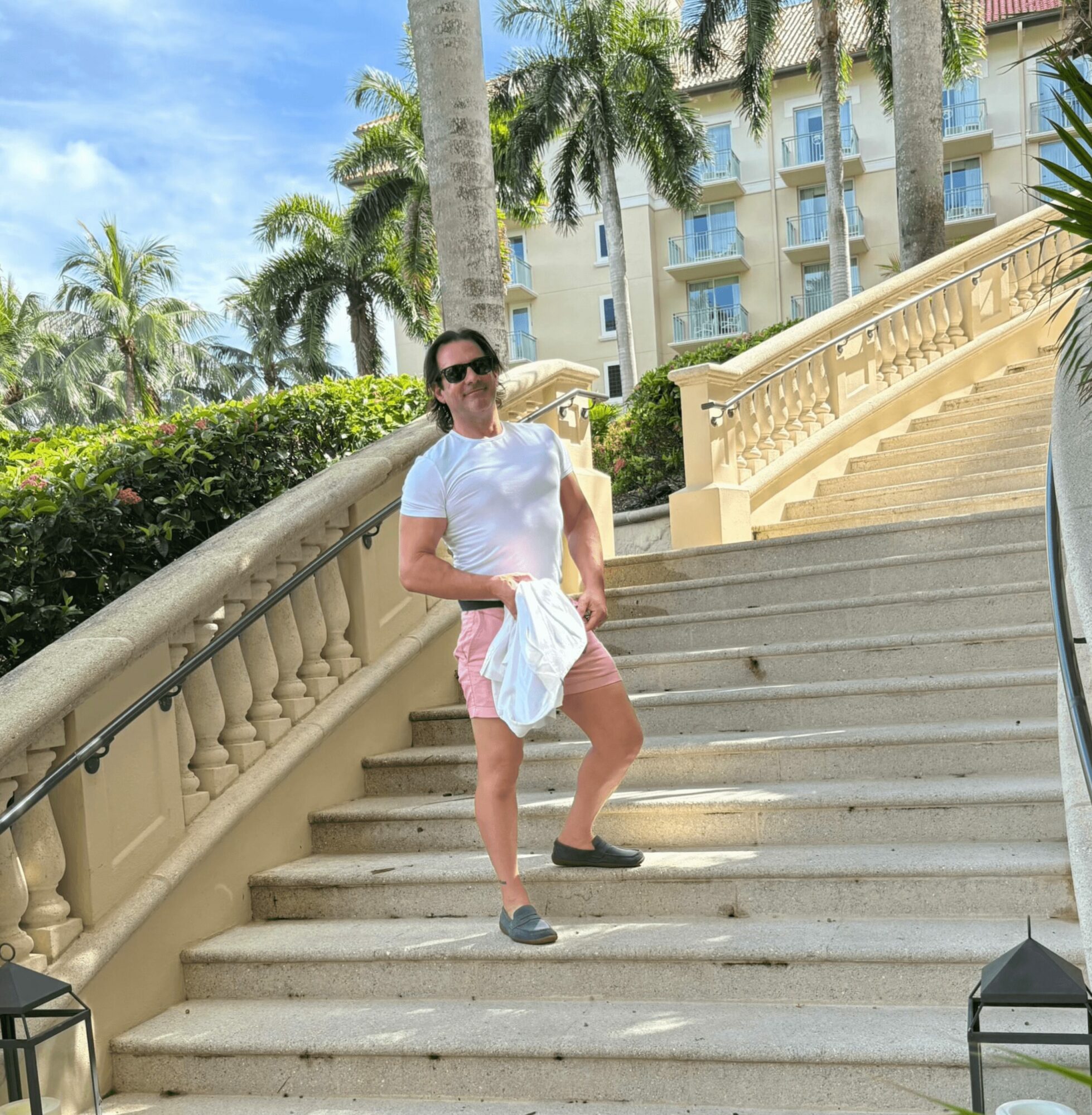 Woman standing on outdoor staircase with palm trees and building in background, holding a white towel, wearing sunglasses, white shirt, pink shorts, and sneakers.