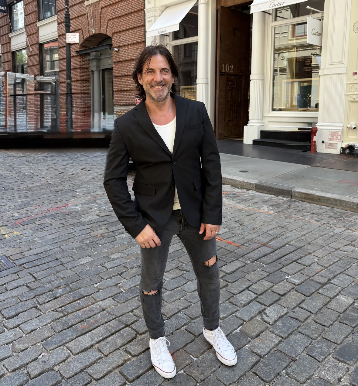Man with dark hair and beard smiling, wearing a black blazer, white shirt, ripped jeans, and white sneakers, standing on cobblestone street.