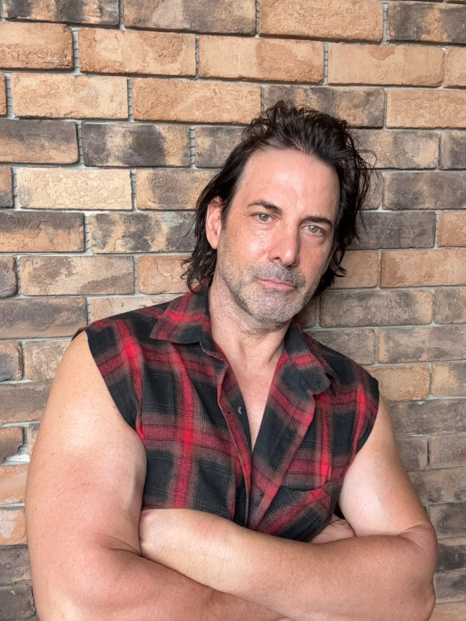Man with dark hair and beard, wearing a sleeveless red plaid shirt, standing against a brick wall.