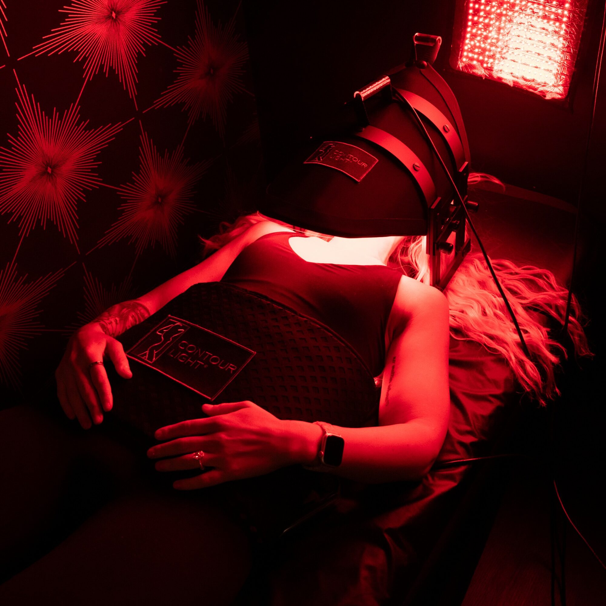 Person lying on bed with a helmet-like device on their head, illuminated by red light, holding a tablet, in a dark room.