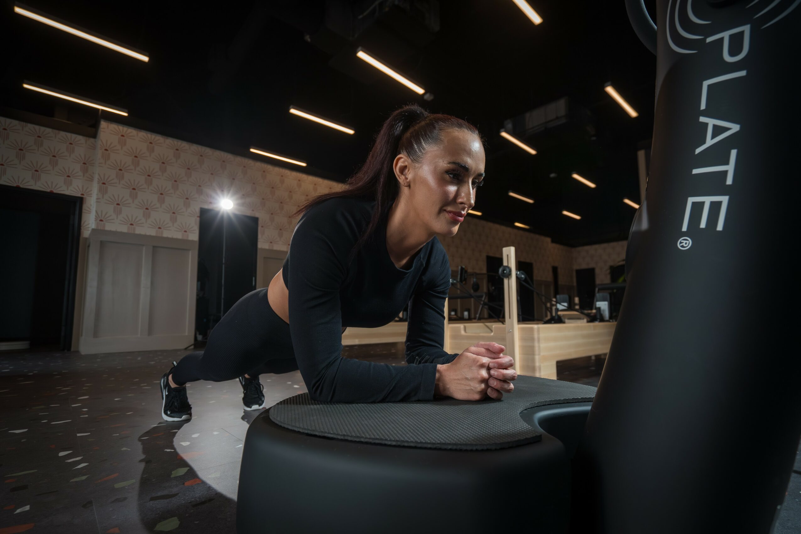 Woman in black workout clothes pushes a punching bag in a gym with black ceiling lights.