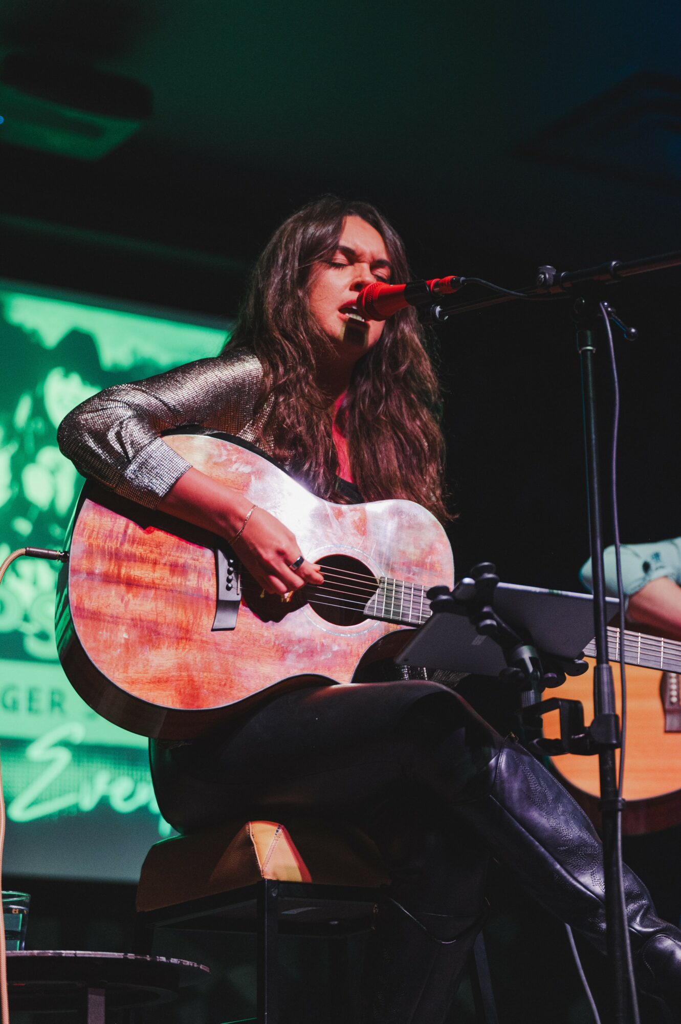 Woman with long dark hair playing an acoustic guitar and singing into a microphone on stage.