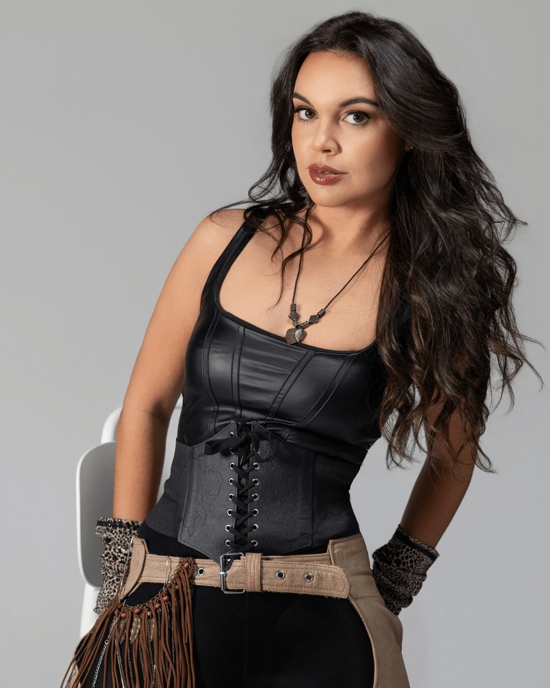 Woman with long dark hair wearing a black top and corset, posing against a gray background.